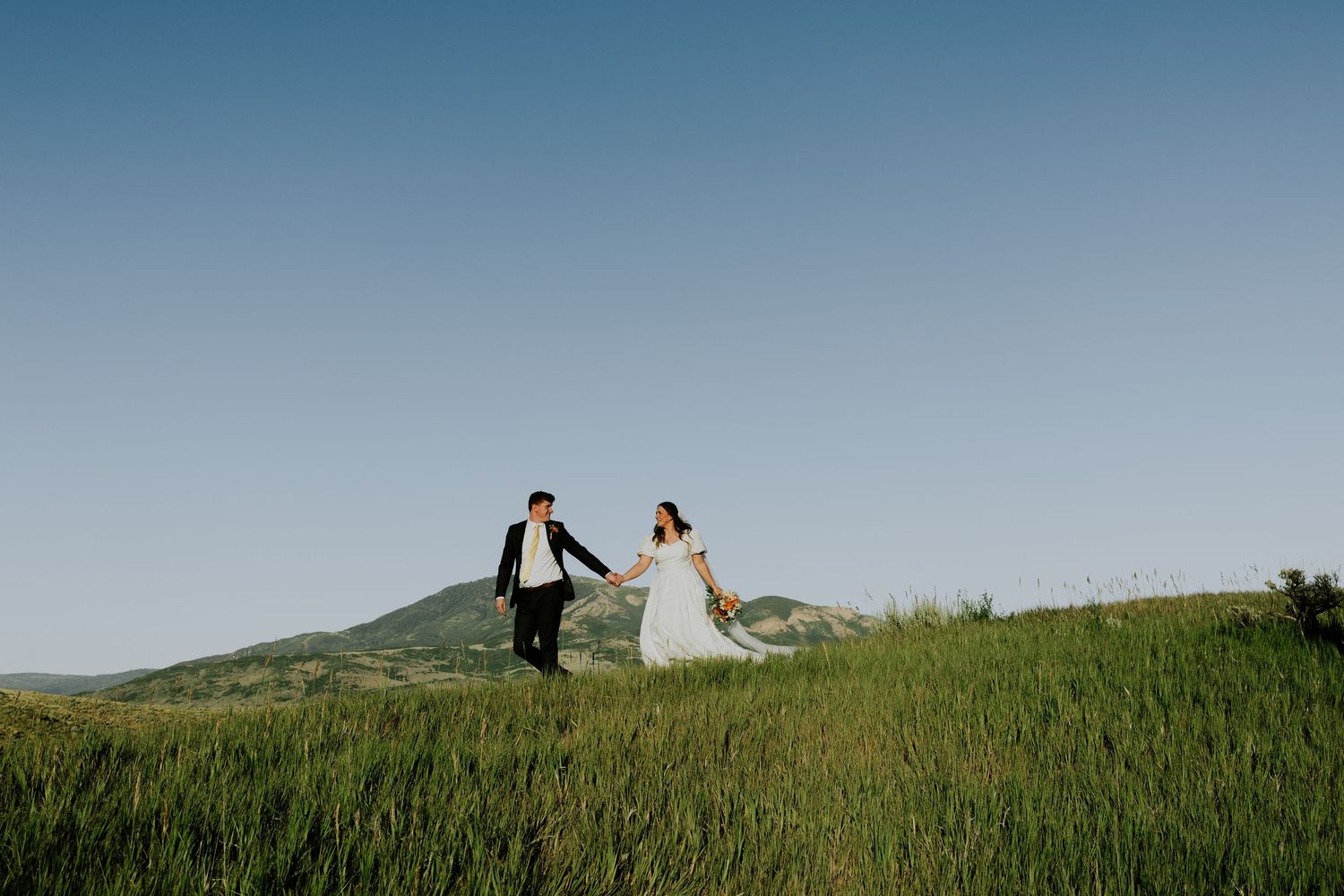 Couple in wedding attire poses together on a scenic green hillside during sunset.