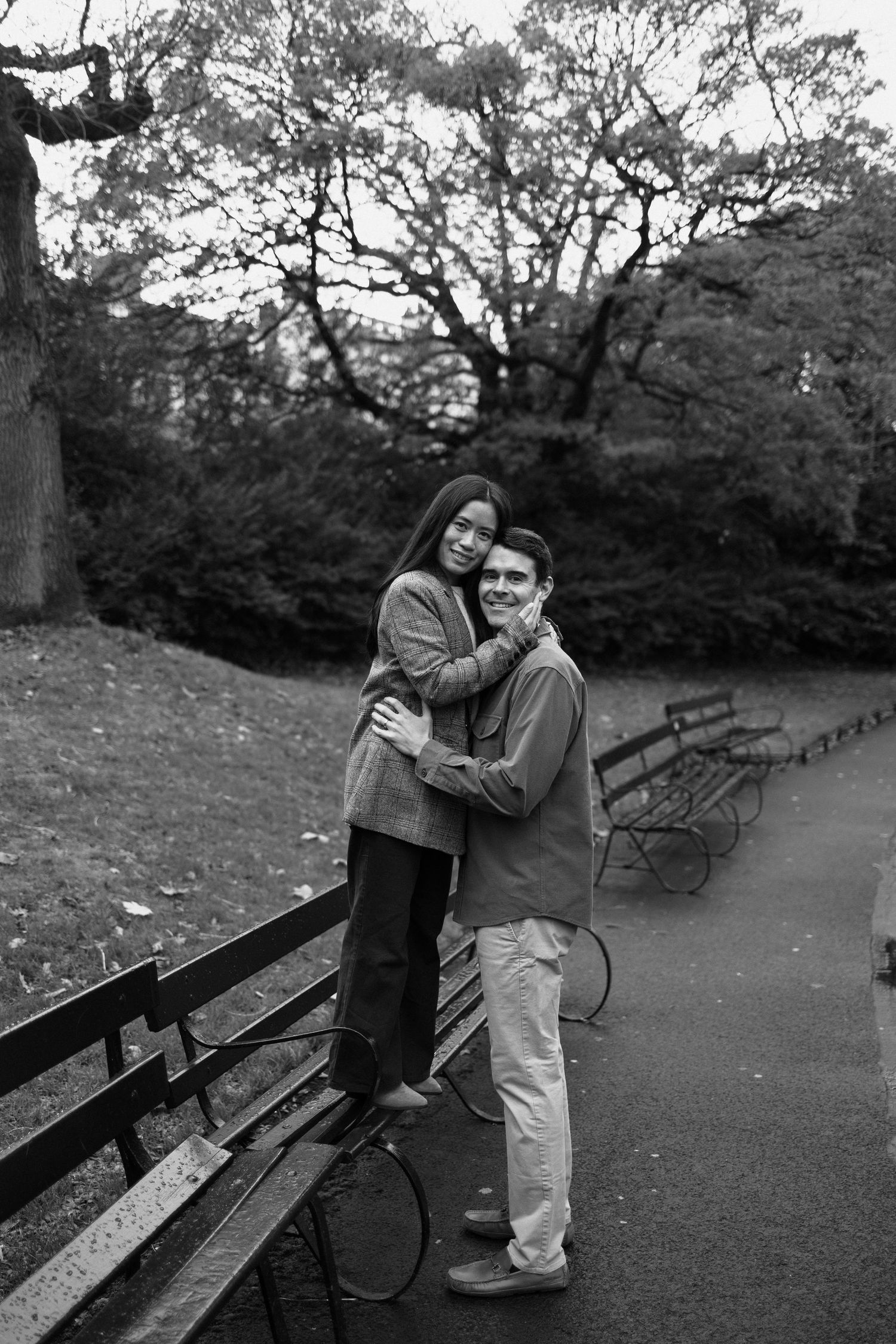 Black and white photo of a couple embracing on a park path surrounded by benches and autumn trees.