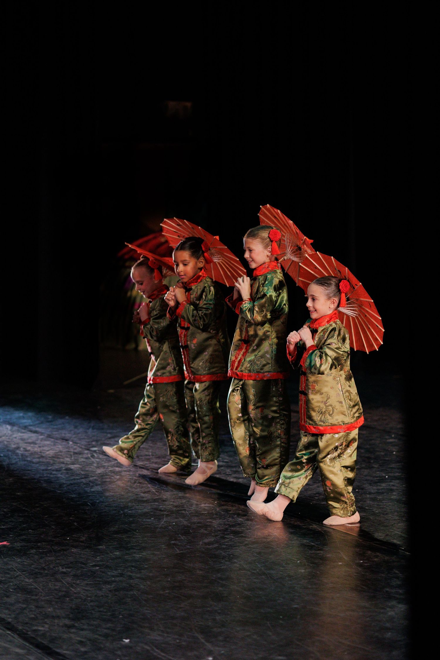 Performers in military uniforms dance with red parasols in a traditional choreographed stage performance.