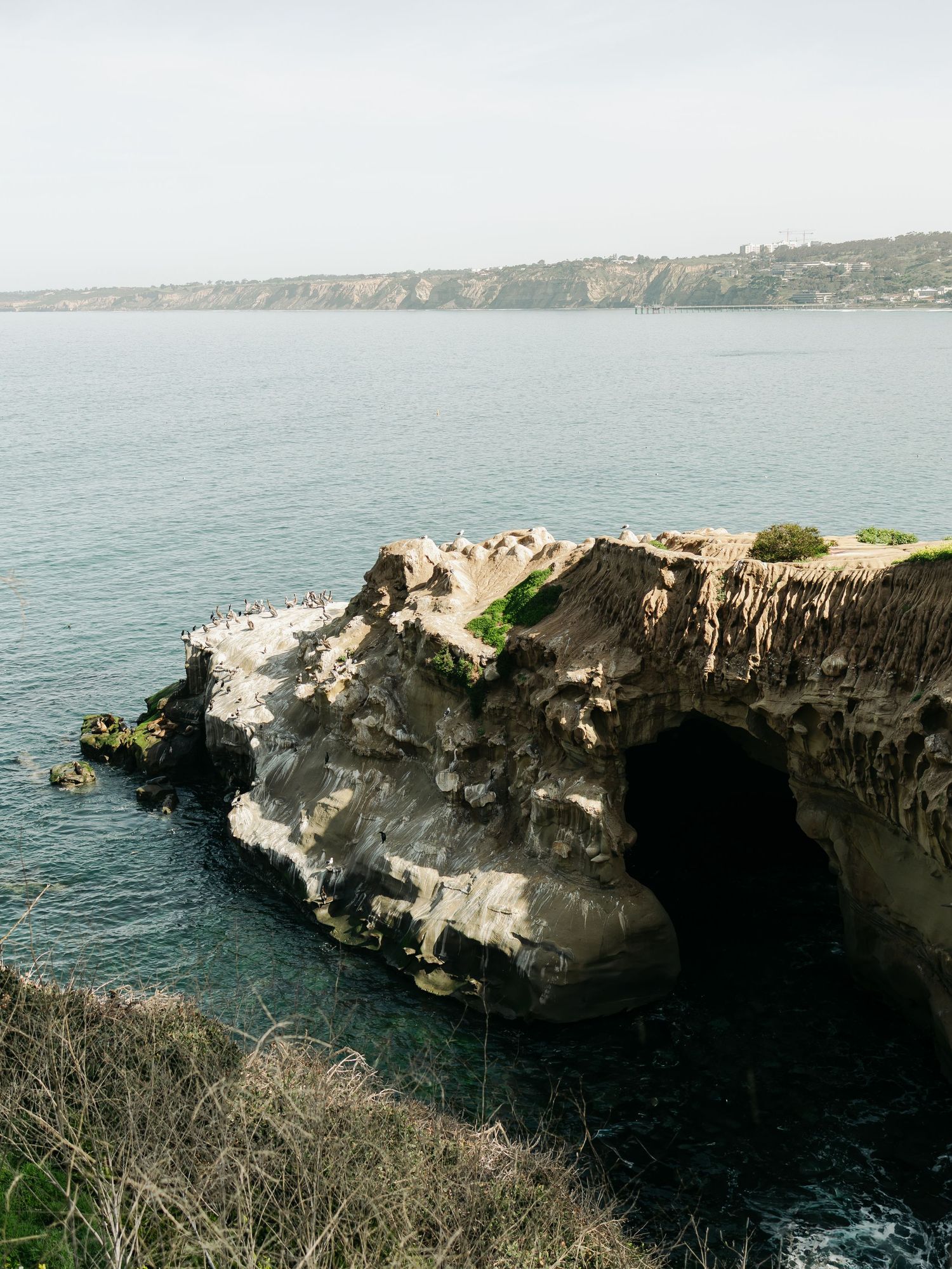 Rocky coastal cliffs with sea cave arch overlooking calm blue ocean waters and distant shoreline under hazy sky.