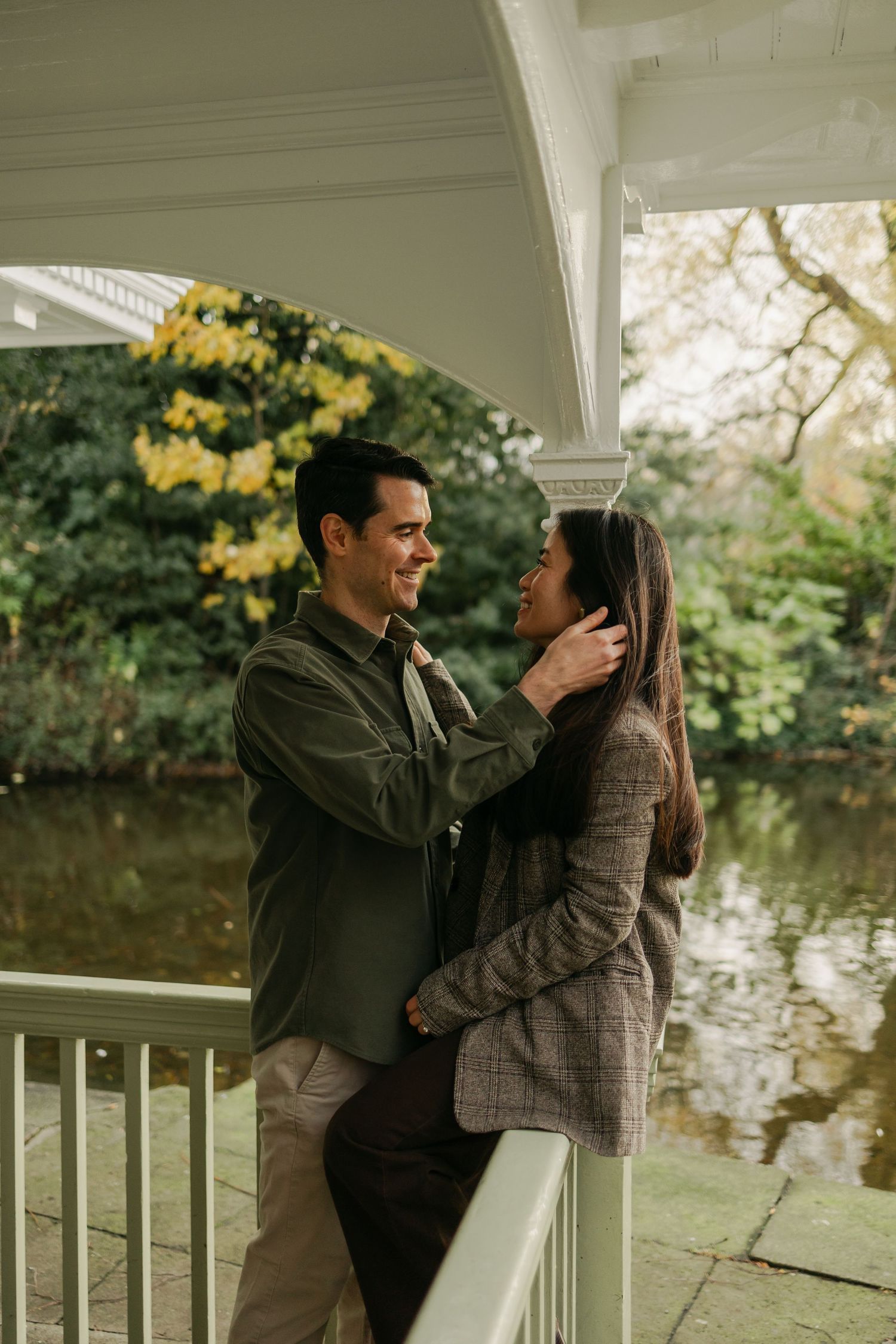Couple sharing a romantic moment under a white gazebo by a pond in a lush green park during autumn.