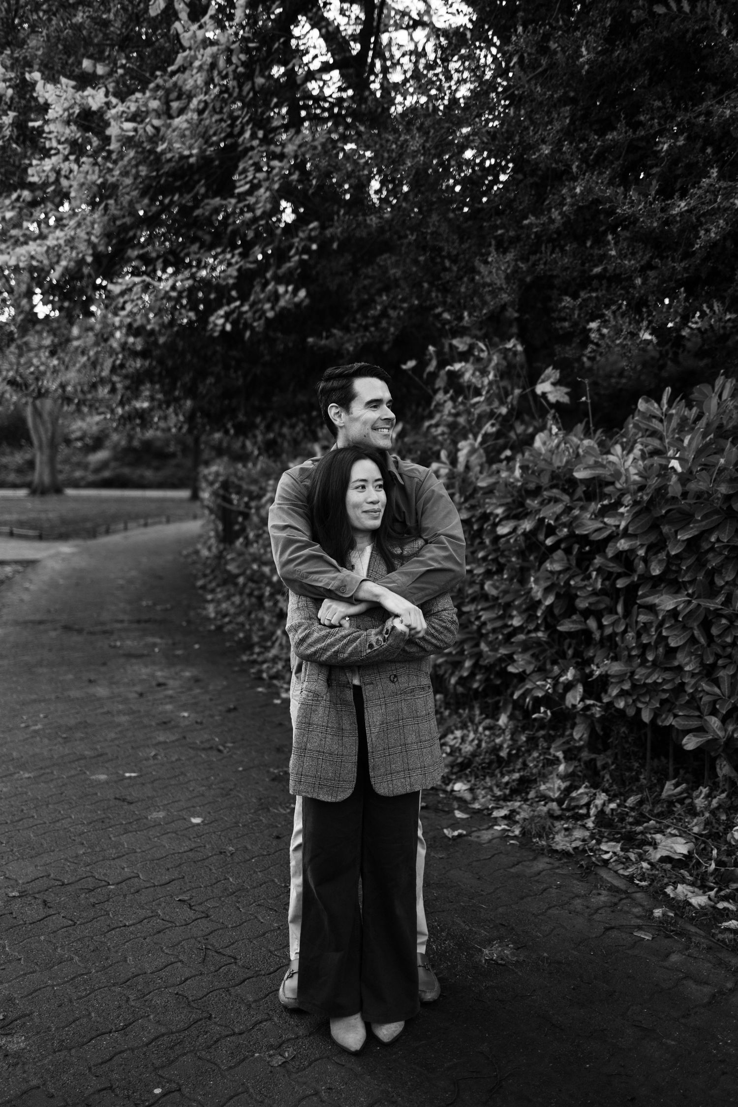 Couple embracing on a park path surrounded by lush greenery, captured in black and white photography.