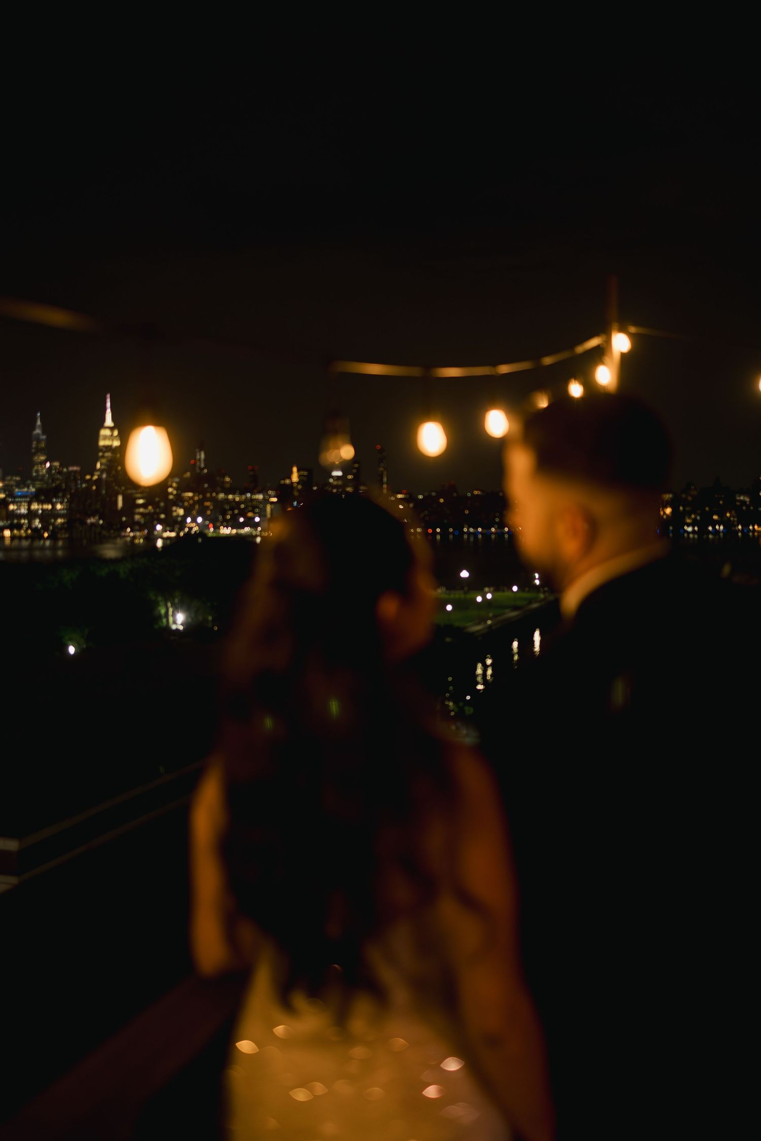 Nighttime celebration on a pier with string lights glowing against city lights across the water.