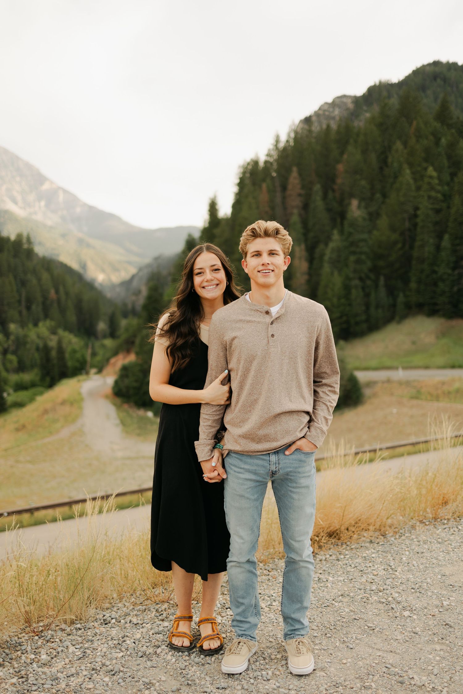 A couple poses playfully on a mountain path surrounded by scenic landscapes.