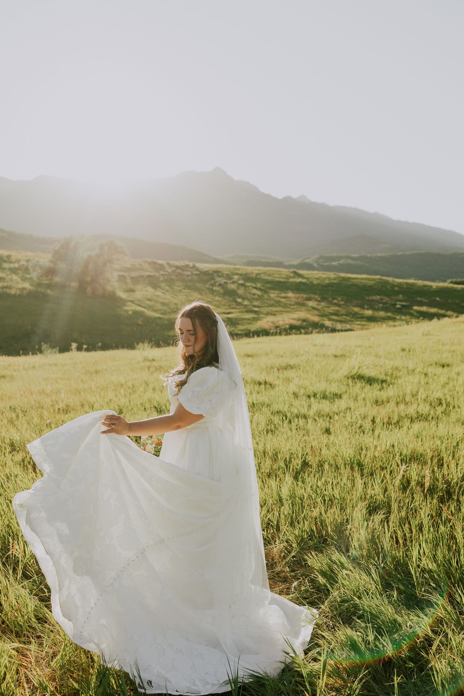 A white wedding dress flows elegantly across a sunlit mountain meadow in a scenic outdoor landscape.