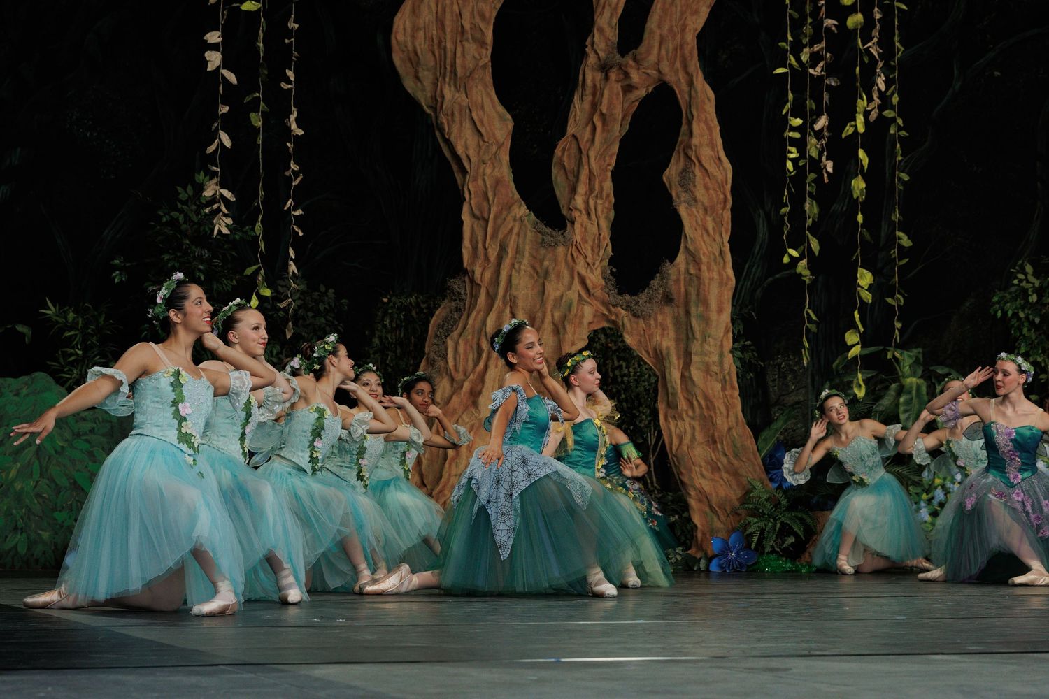 Corps de ballet dancers in flowing aqua tutus perform in unison before a large tree backdrop on stage.