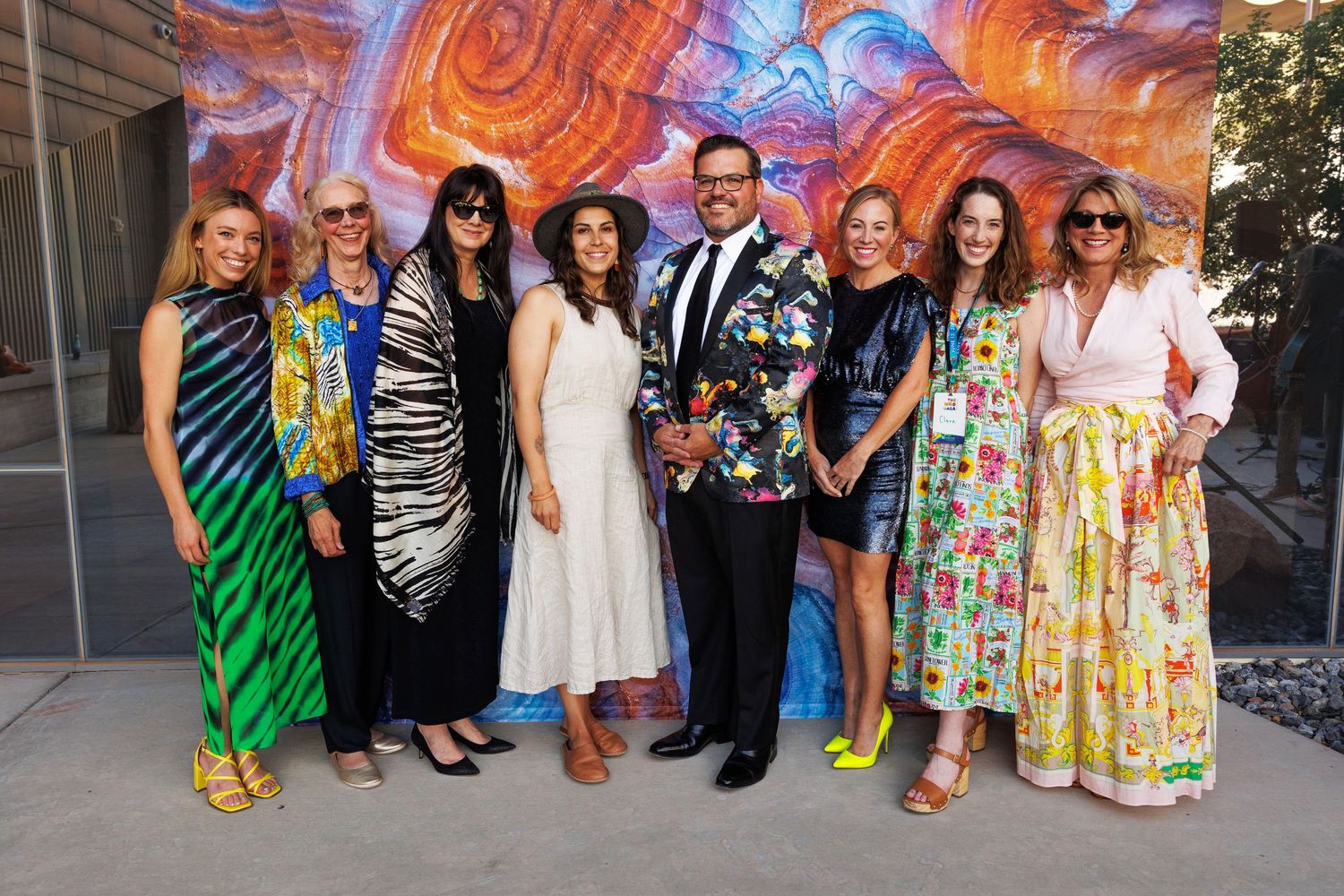 Group of eight people posing together at a colorful art event, dressed in vibrant outfits in front of a mural.