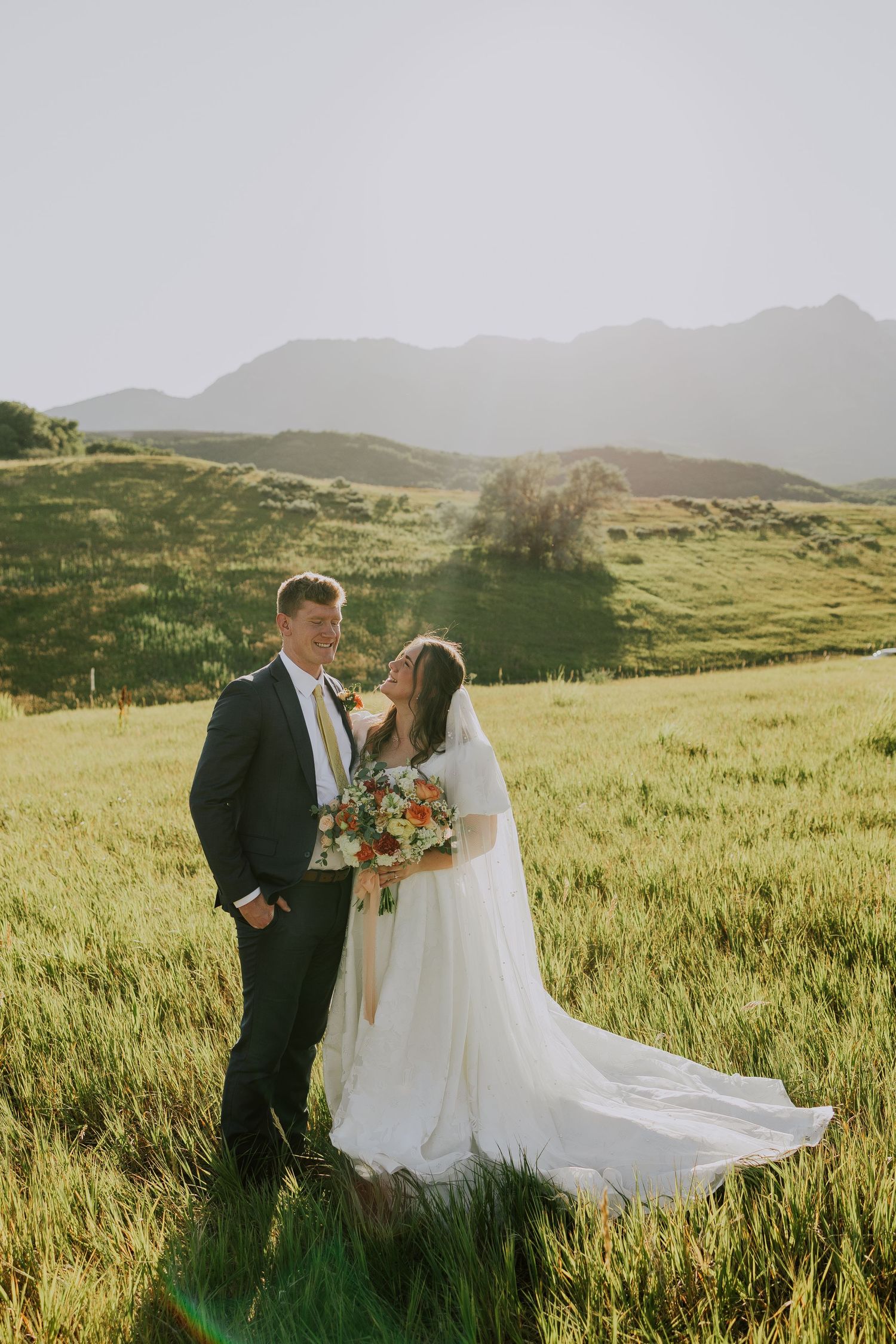 A wedding couple stands in a sunlit meadow with rolling green hills and mountains in the scenic background.