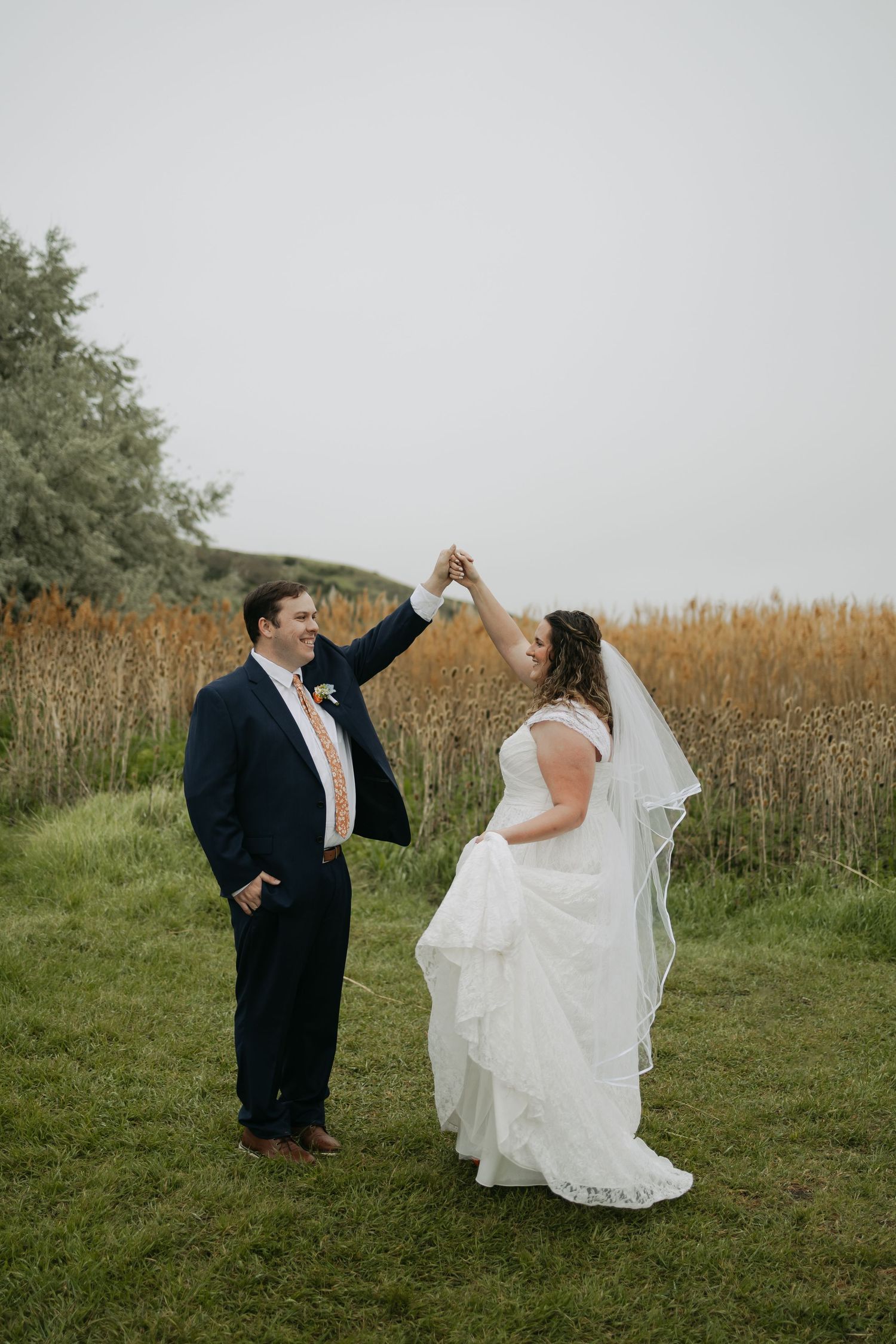 Bride and groom dancing outdoors on grass near golden reeds, groom twirling bride in white gown and veil on cloudy day.