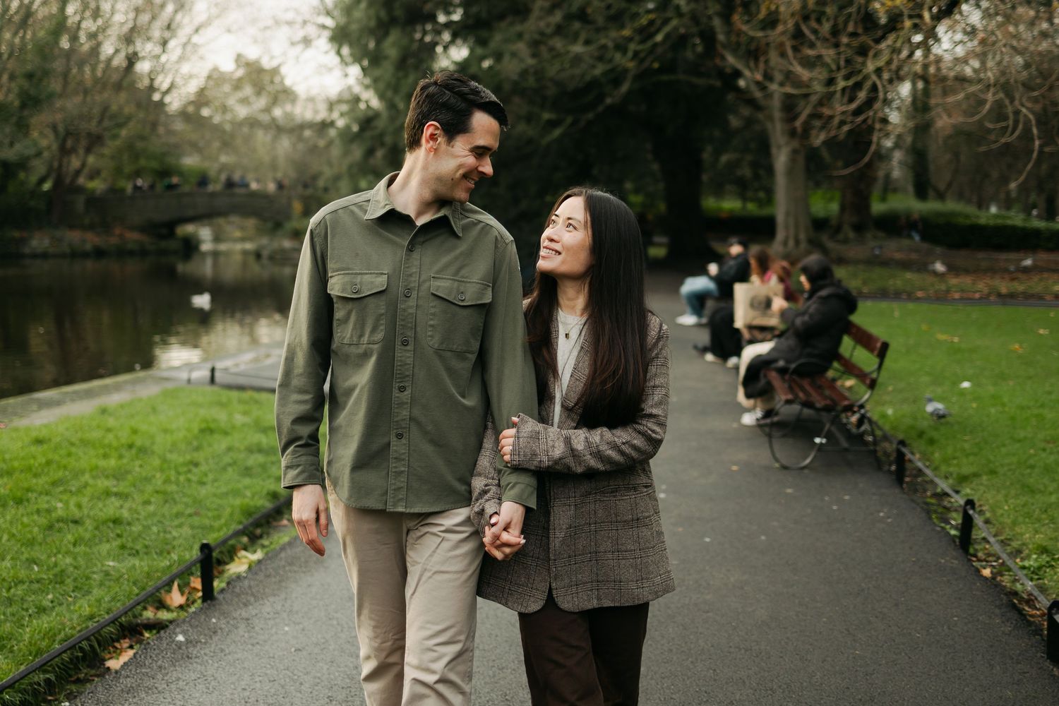 A couple walks hand in hand through a lush green park, smiling at each other on a paved pathway.
