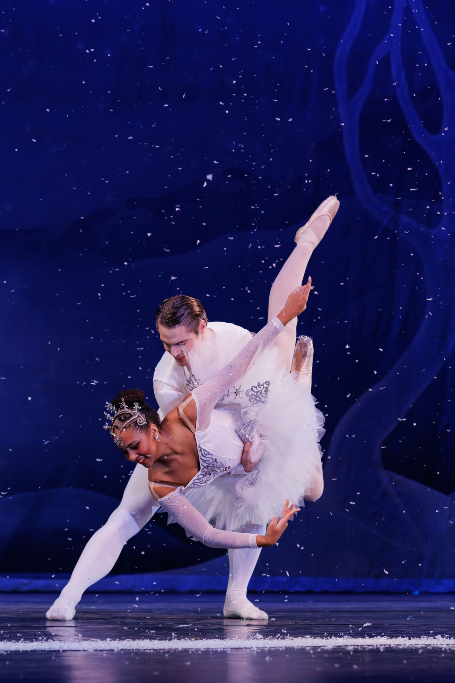 Ballet dancers perform a graceful sequence of sliding movements across an icy stage with a dark blue backdrop.