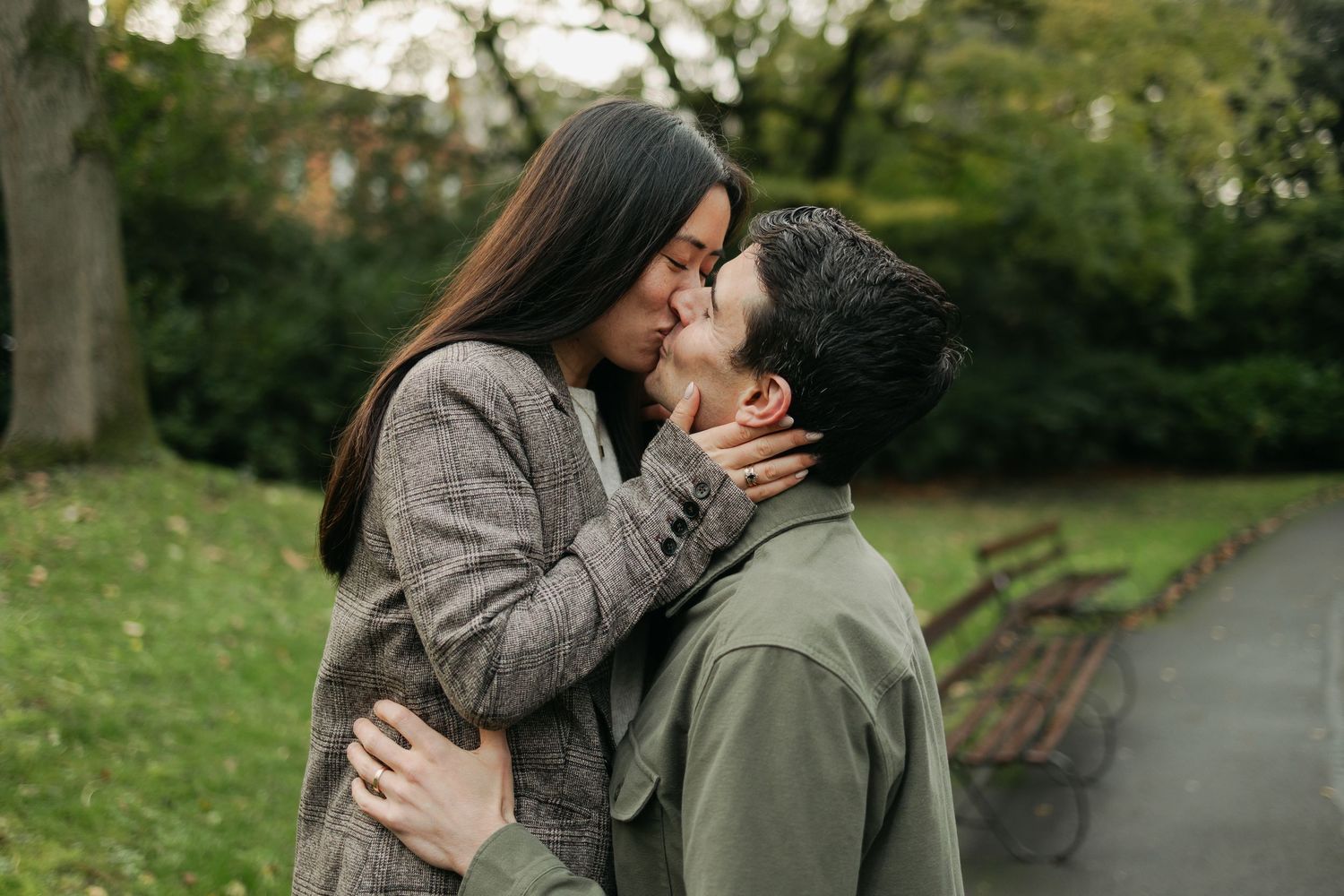 Couple sharing a romantic kiss in a park during autumn, surrounded by green grass and trees.