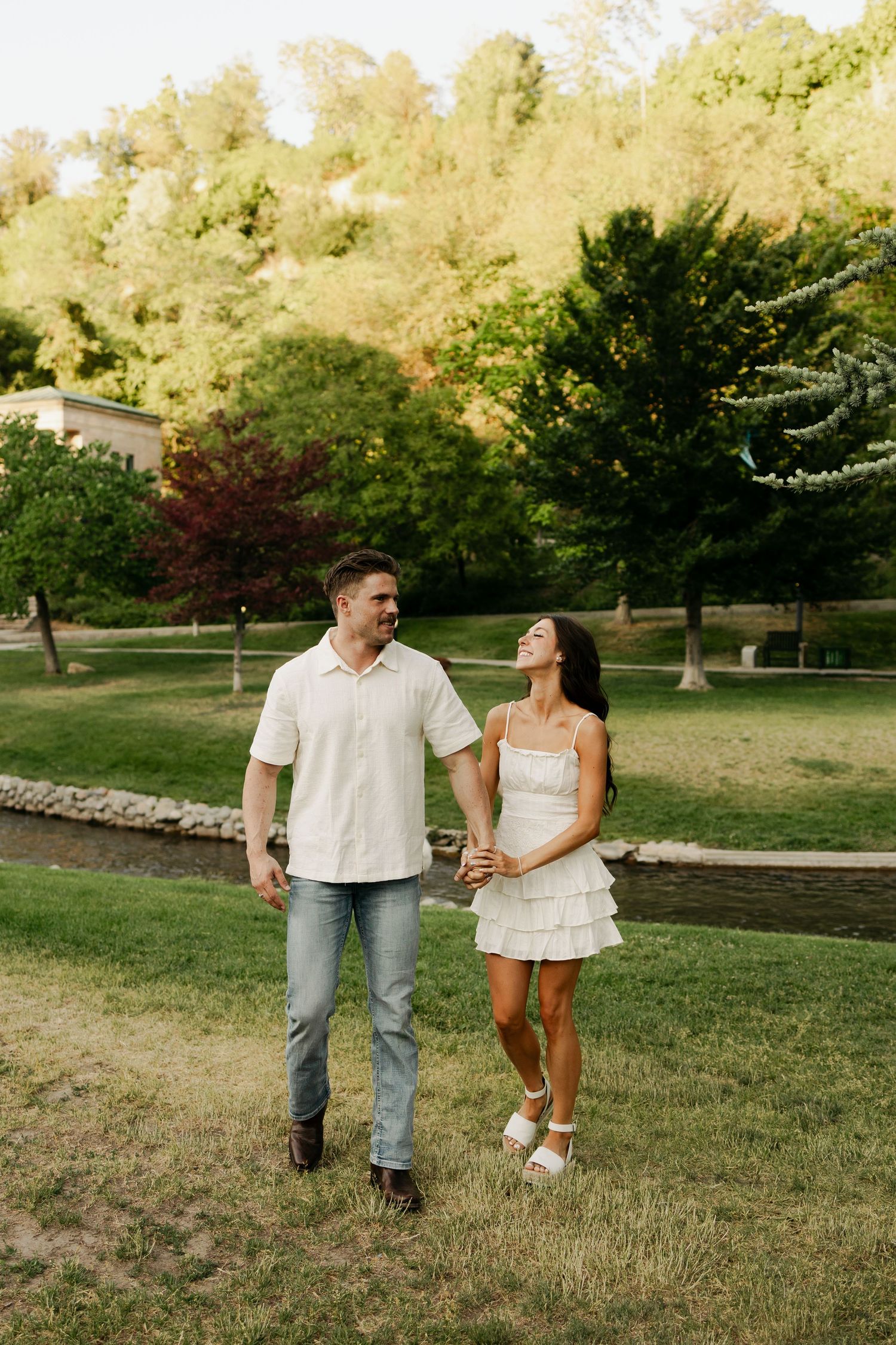A couple walks and laughs together on a lush green lawn surrounded by trees on a sunny summer day.