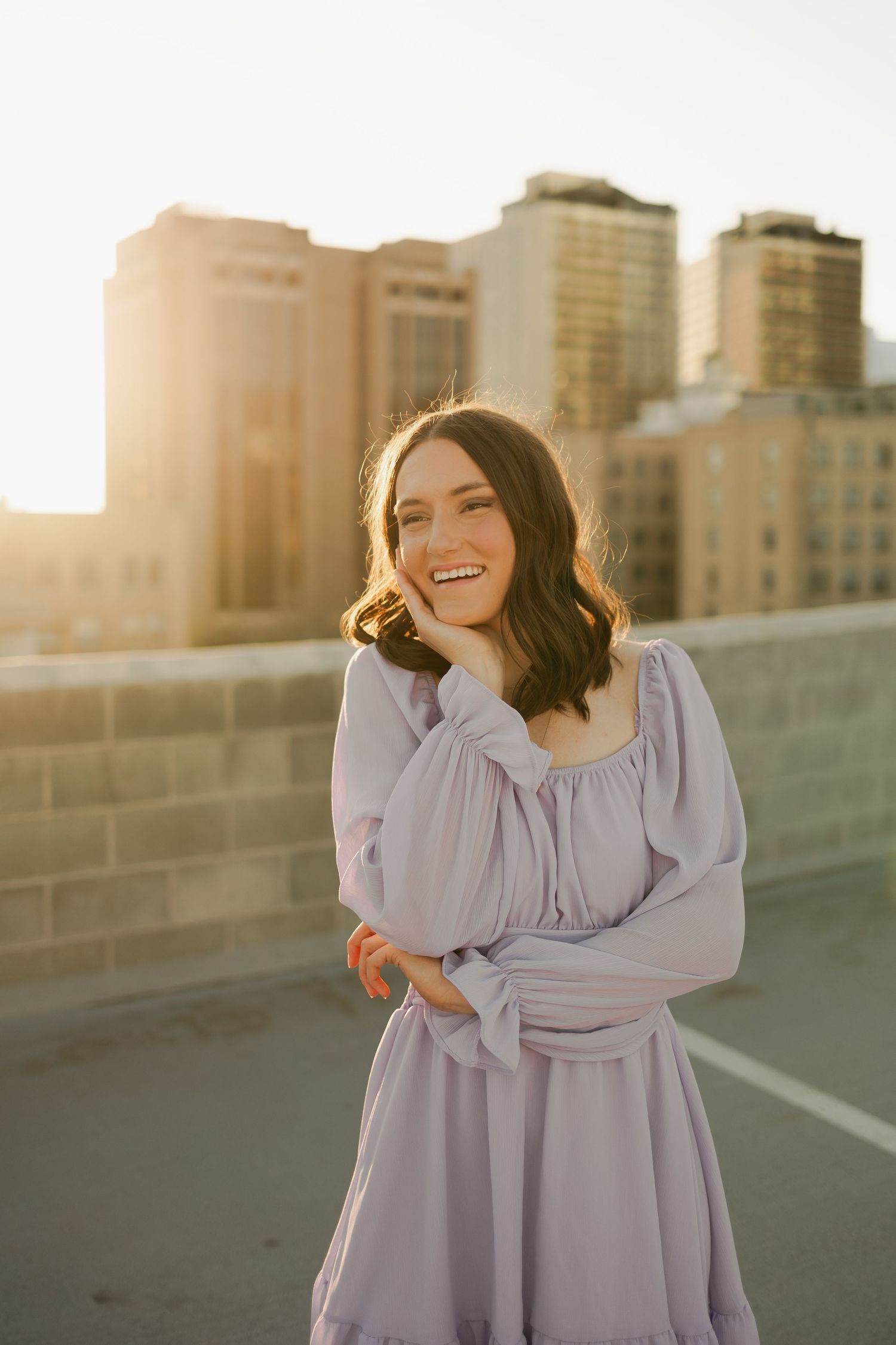 Figure in lavender dress poses against urban skyline at golden hour.