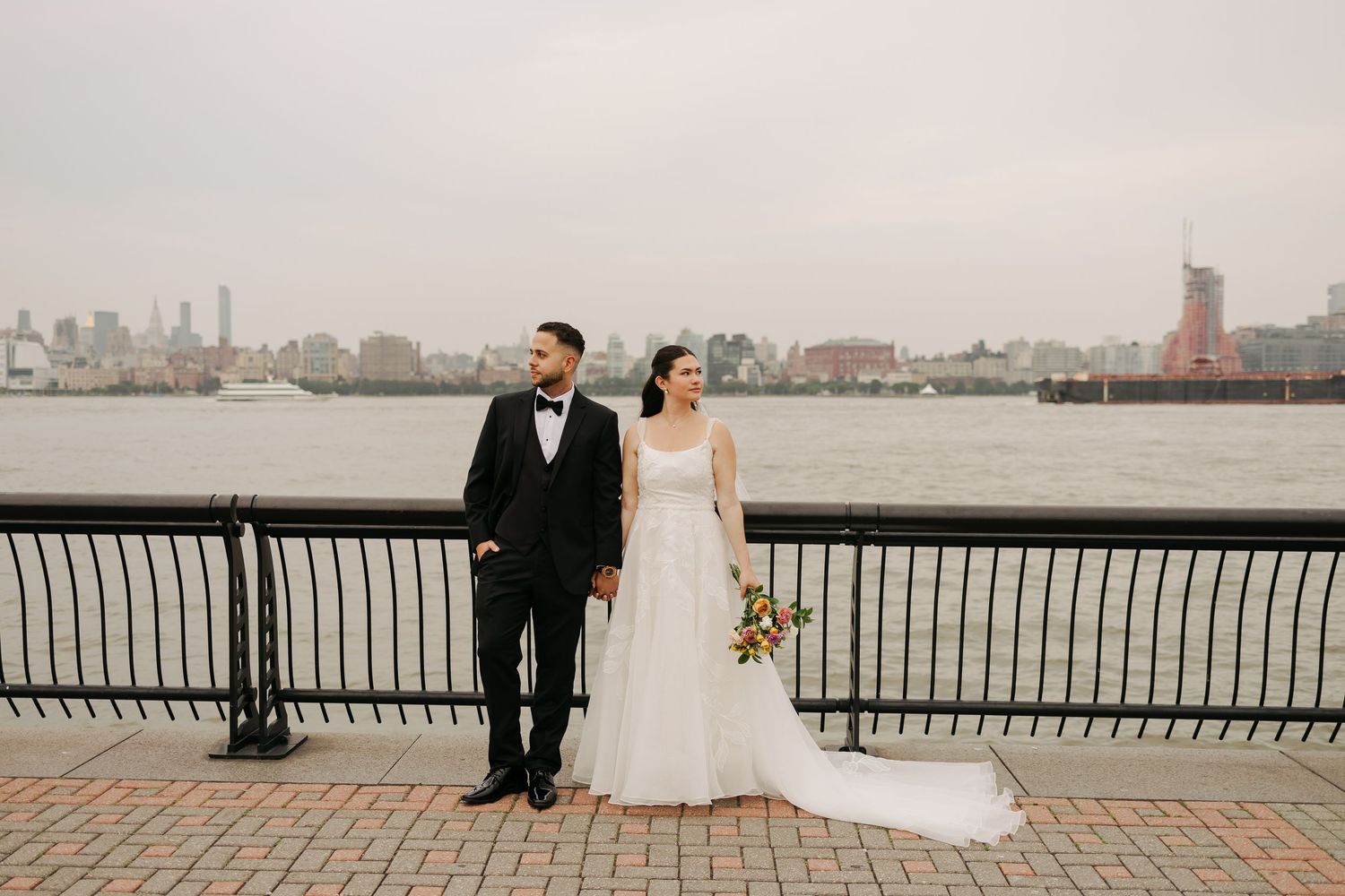 A wedding couple stands by a waterfront railing with city skyline in the background.