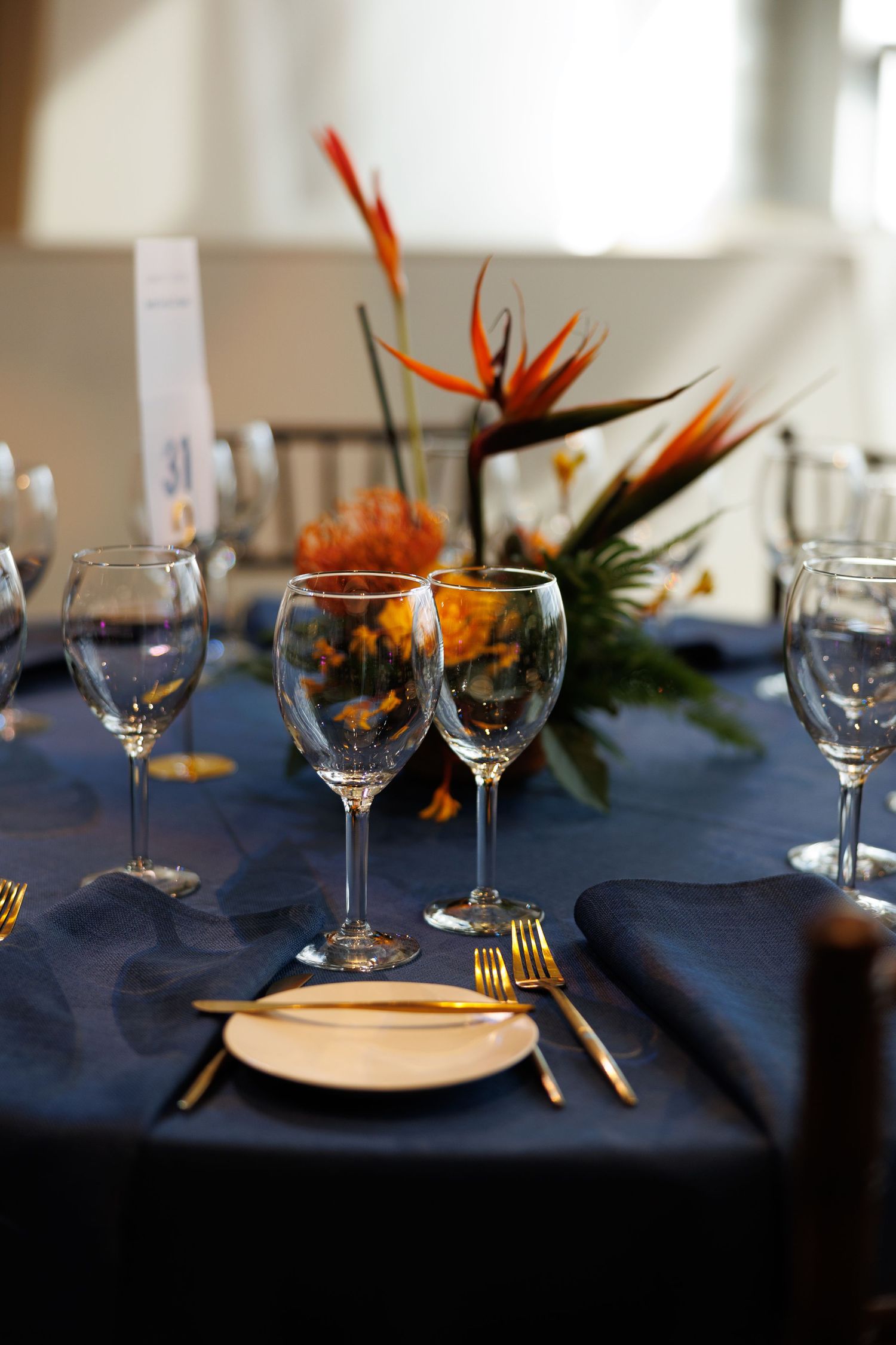 Elegant navy blue table setting with gold cutlery, crystal wine glasses, and bird of paradise floral centerpiece.