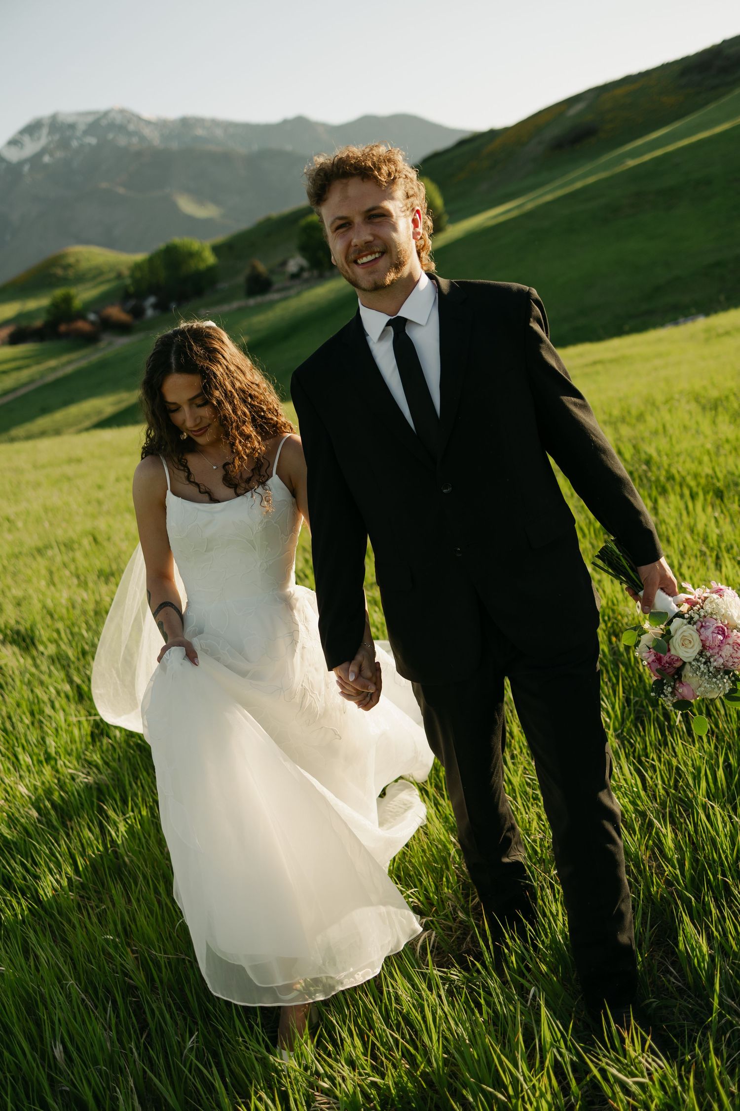 A wedding couple walking through a lush green meadow with mountain peaks in the background.