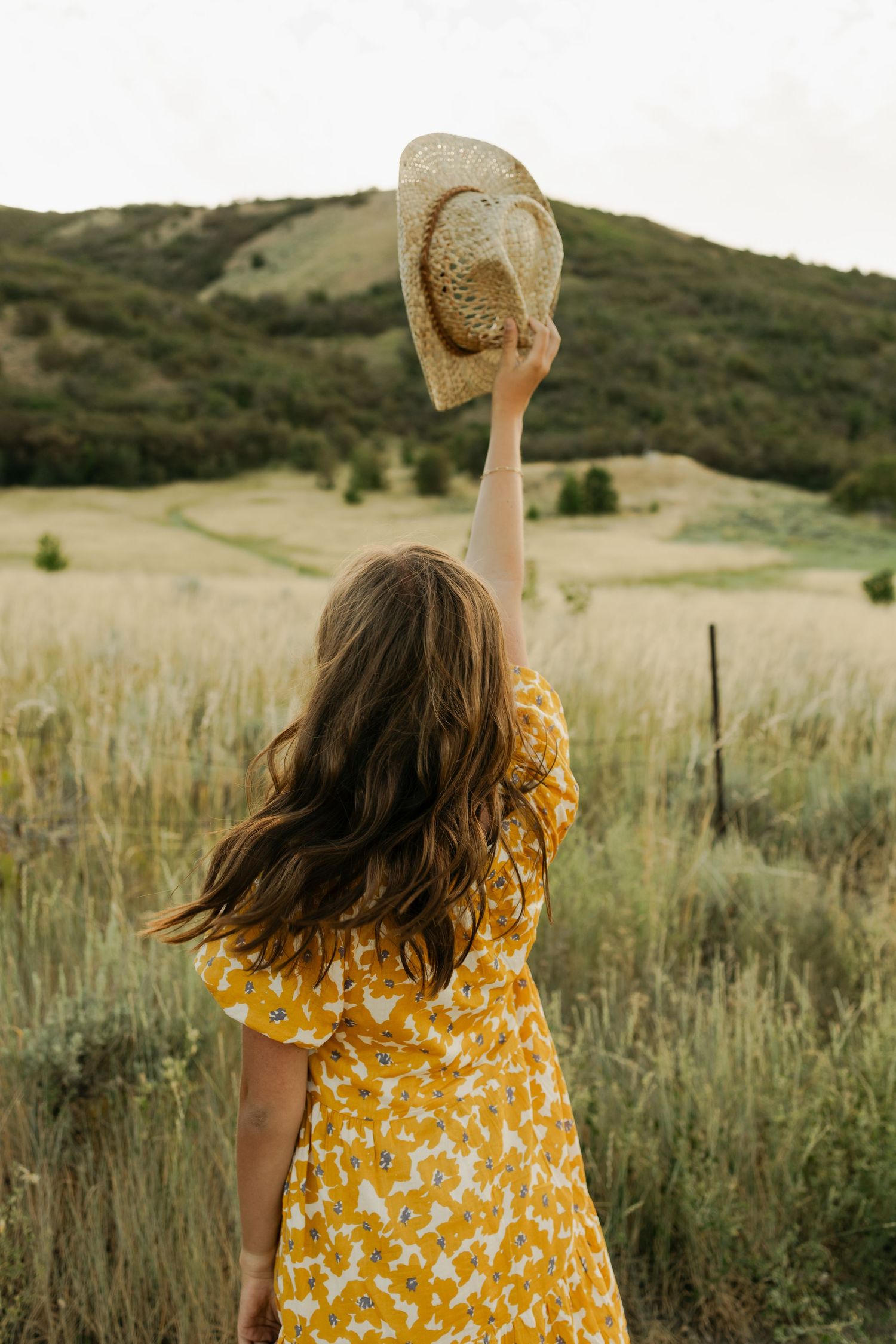 Figure in yellow floral dress holds straw hat while walking through tall grass meadow.