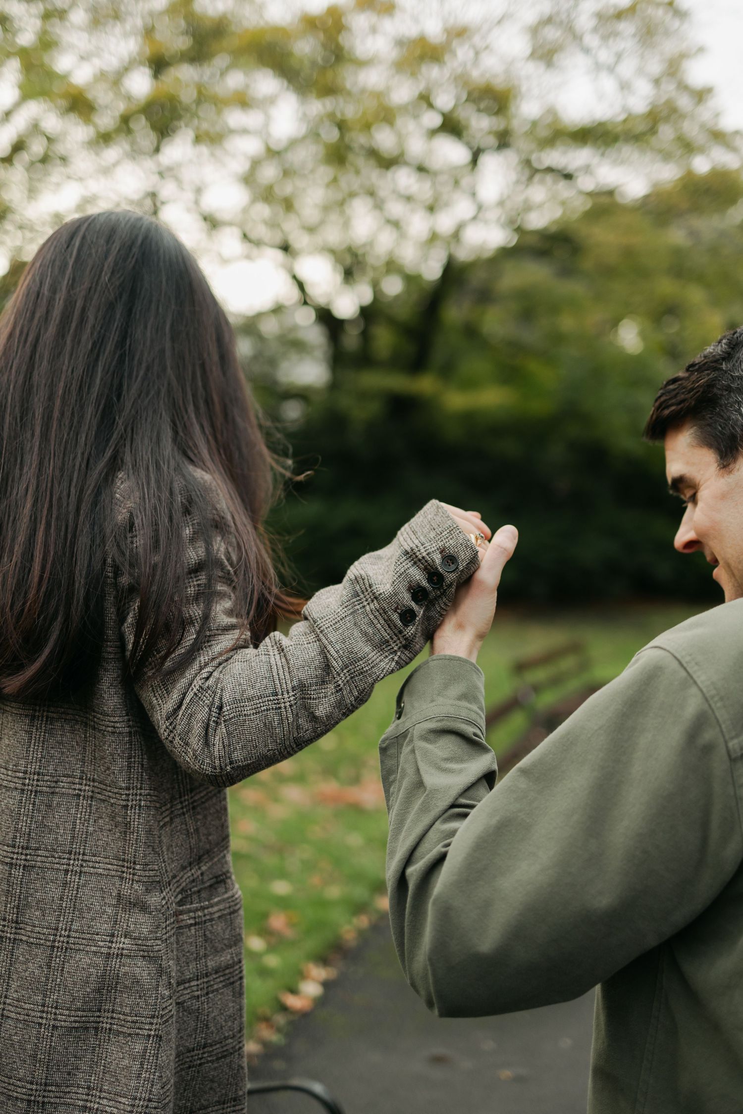 Couple holding hands in a park, man in green jacket and woman in plaid blazer, autumn trees behind.