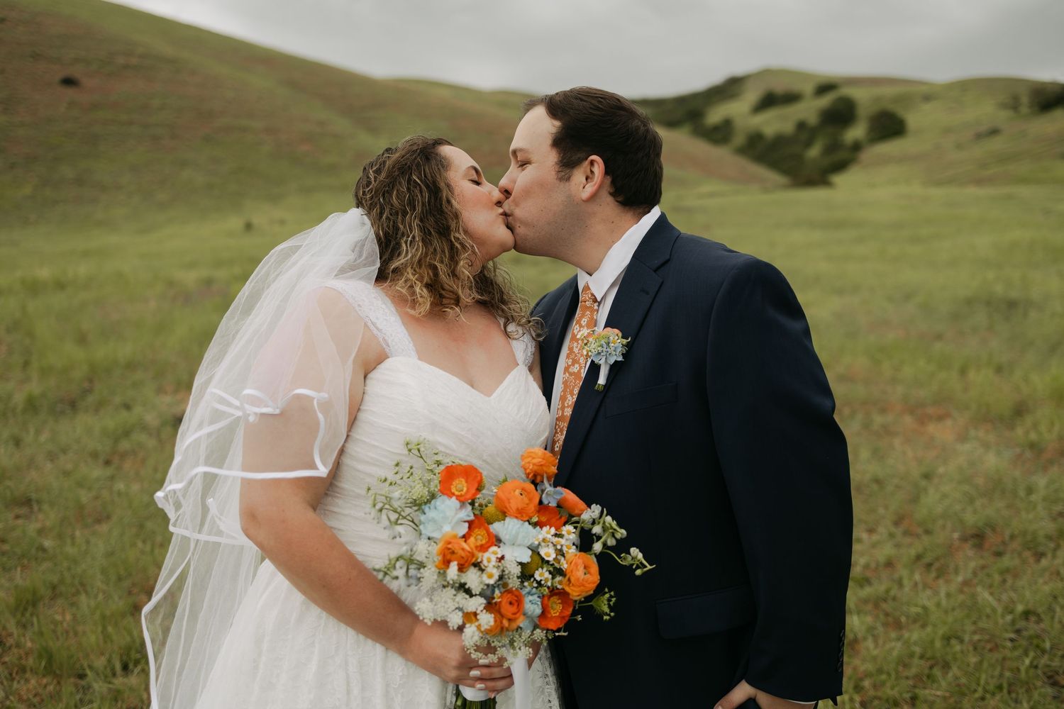 Bride and groom share a kiss in a green meadow, bride holding vibrant orange and blue bouquet wearing white gown and veil.