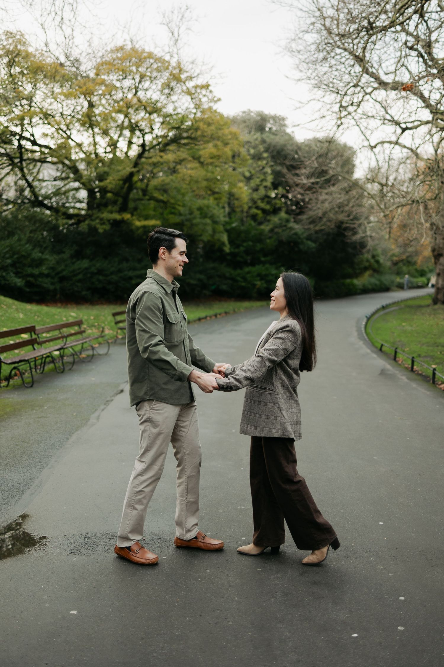 Couple holding hands and smiling at each other on a park path surrounded by lush green trees.