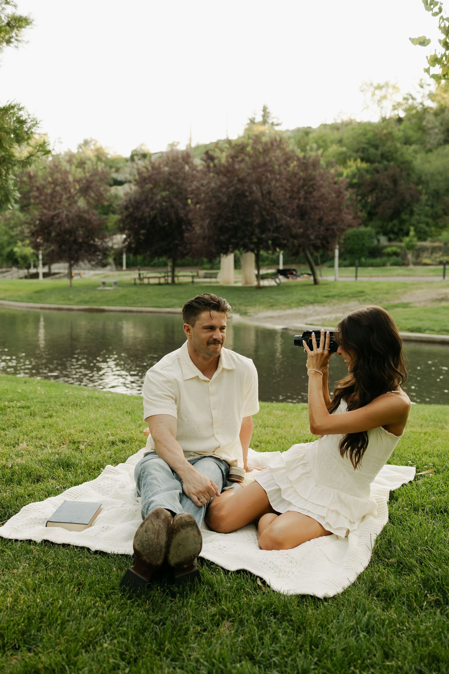 A couple enjoys a playful moment together while relaxing on a white blanket by a lake on a sunny afternoon.