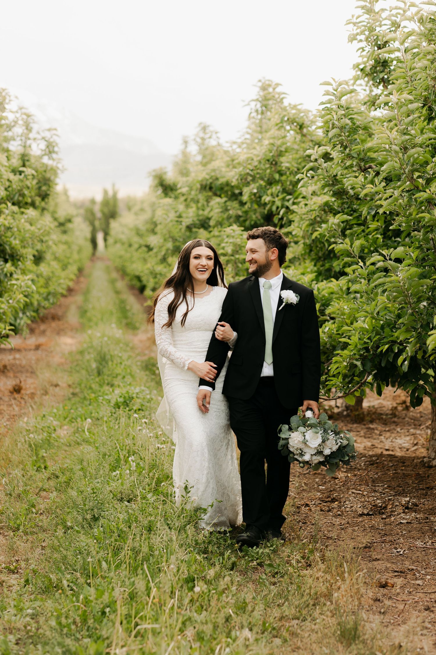 Newlywed couple walking through orchard rows in formal wedding attire.