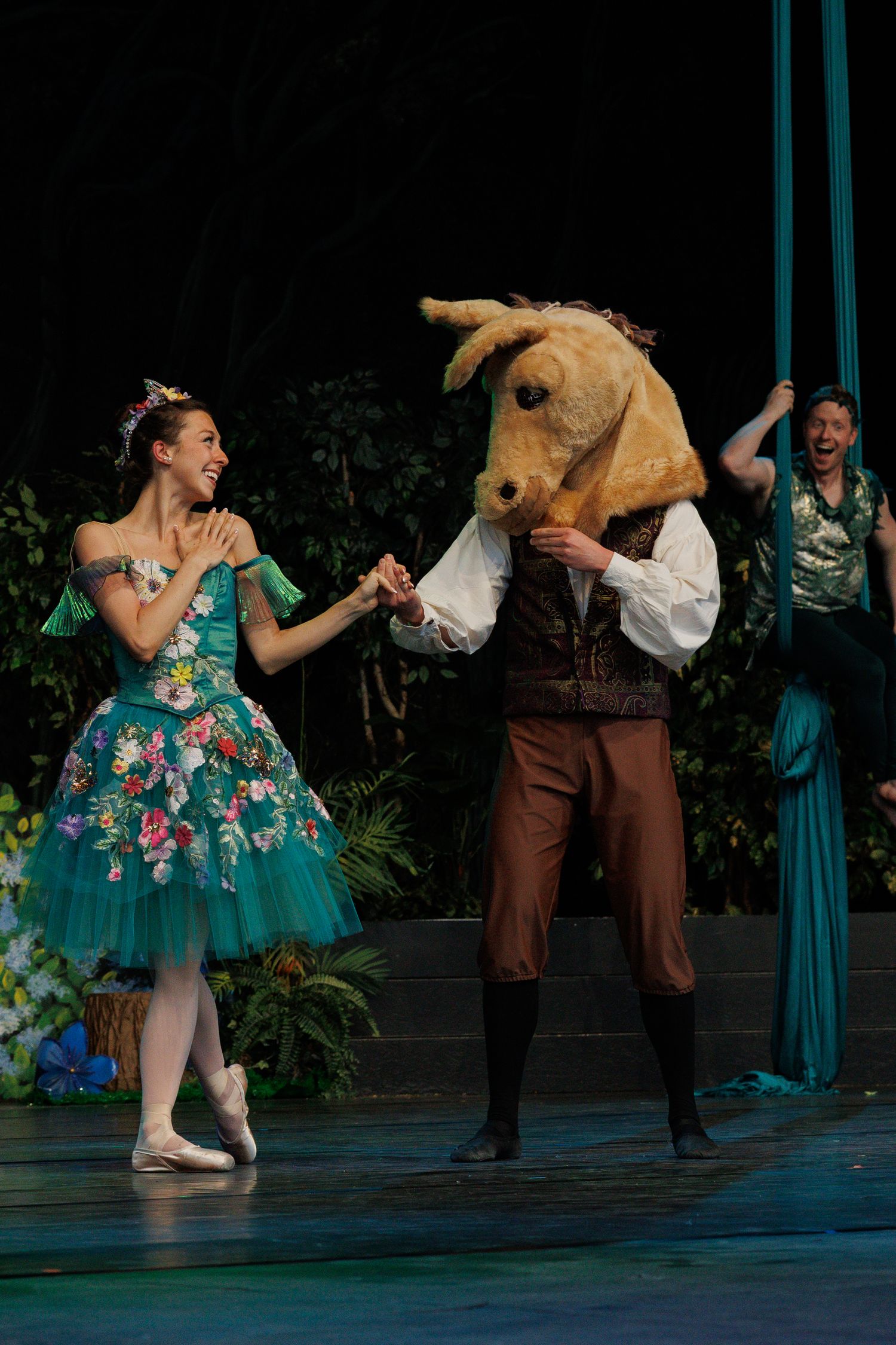 Ballet dancers perform partnered dance scene with turquoise and floral costumes on dimly lit stage.