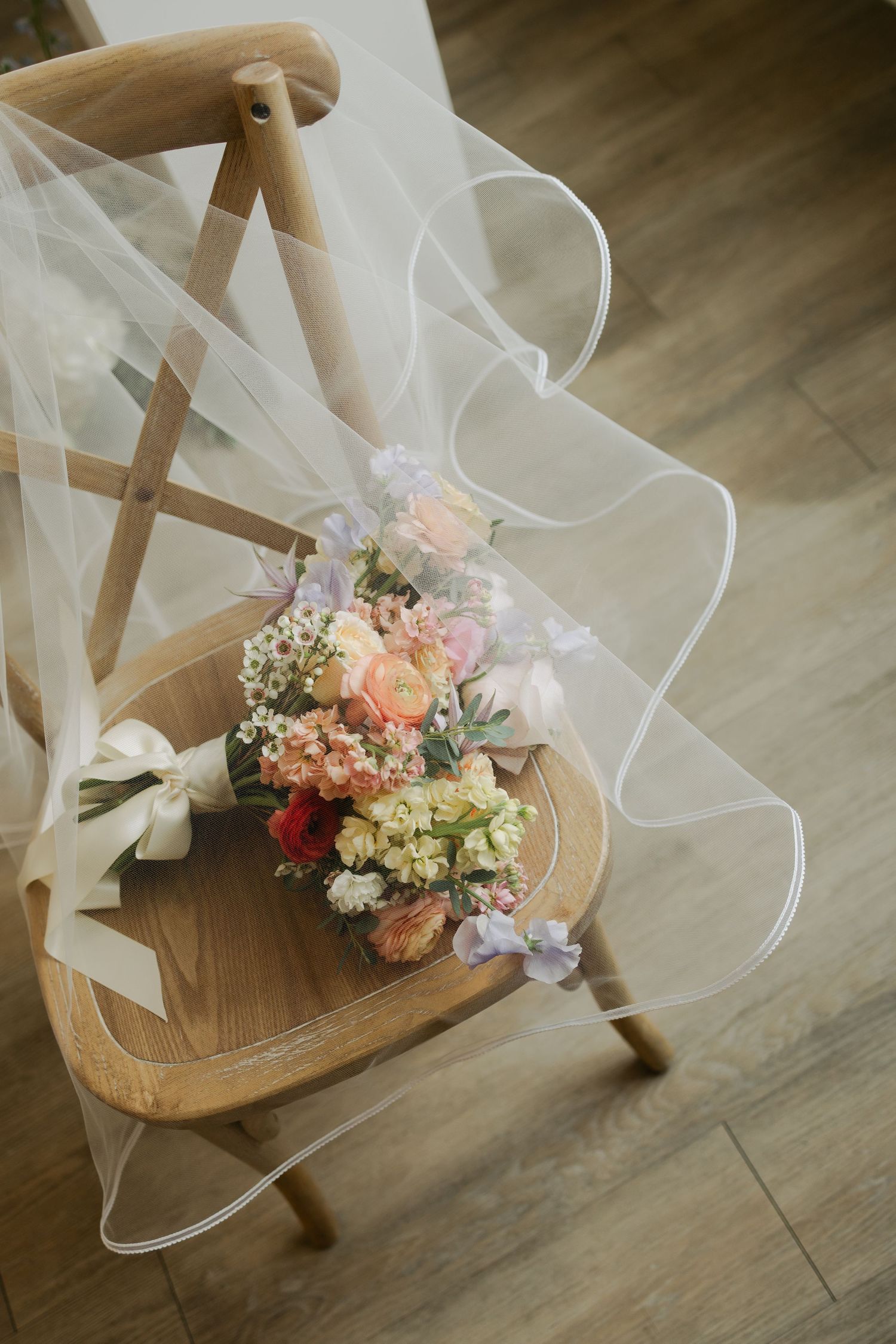 A delicate floral bouquet with white and peach flowers rests on a wooden chair against a softly lit background.