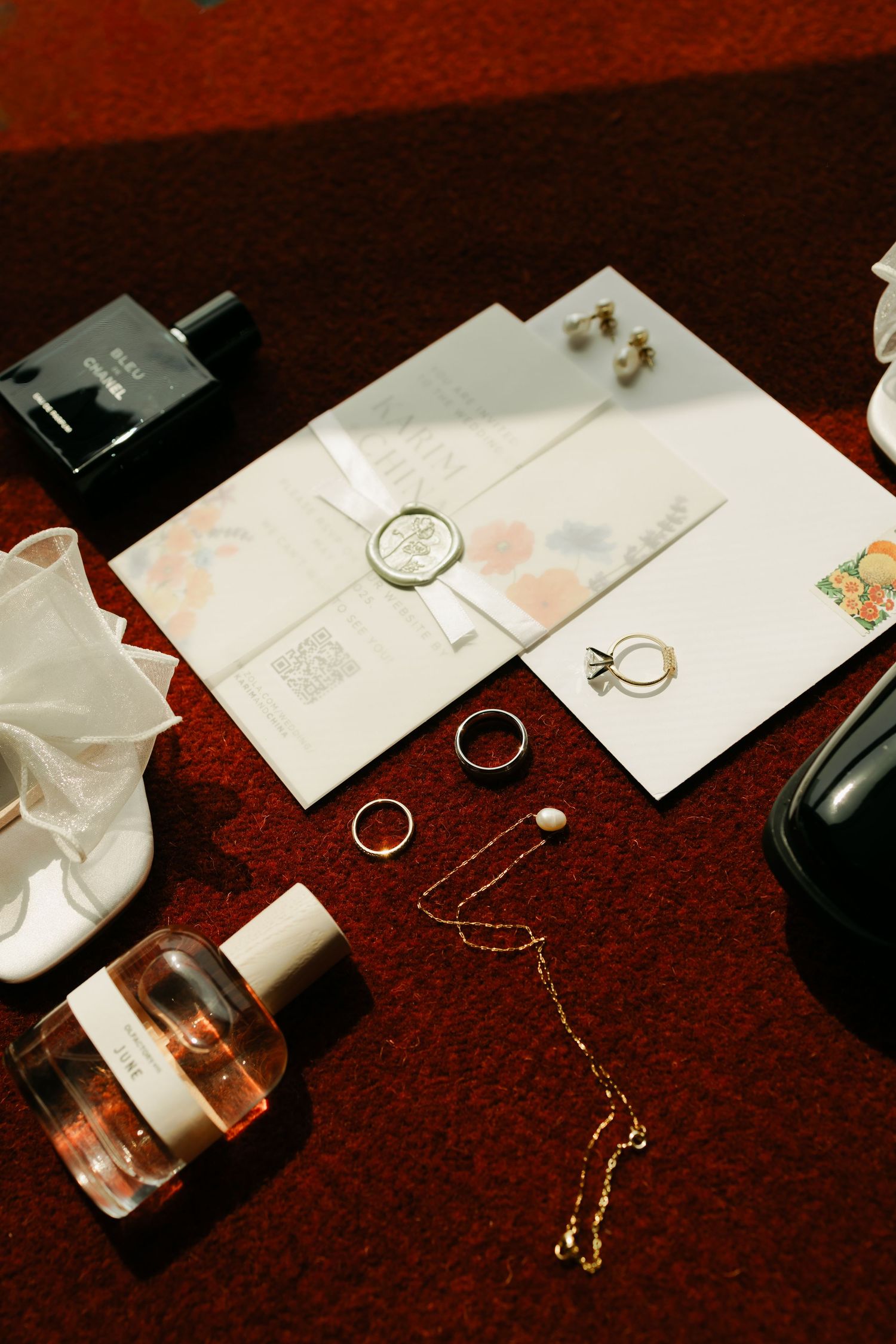 Wedding details including rings, invitations, perfume bottles and jewelry arranged on a deep red surface.