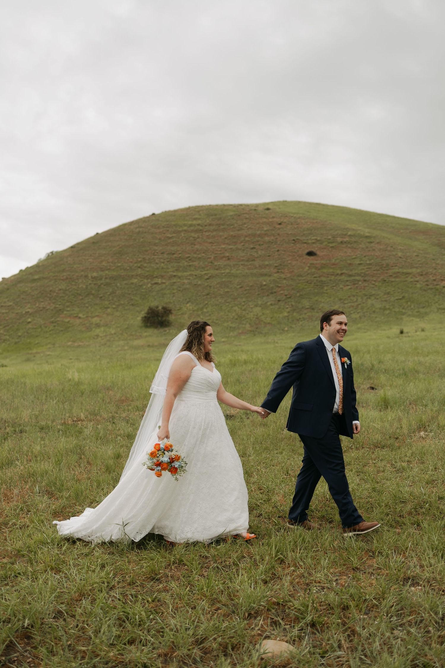 Bride and groom walk hand-in-hand through green meadow with rolling hill backdrop on their wedding day.