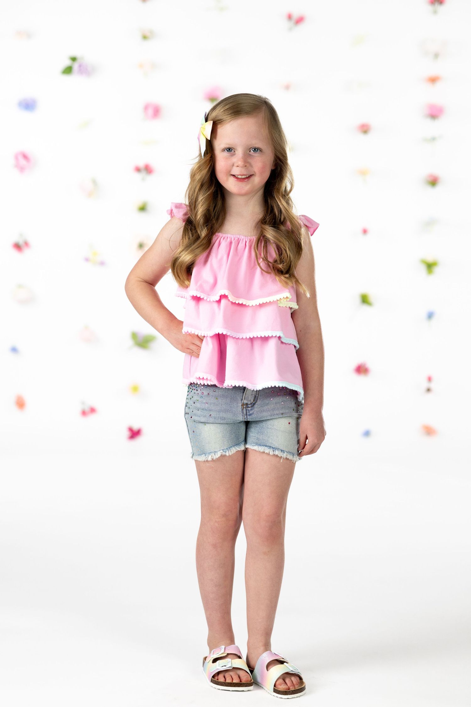 Child wearing pink ruffle top and denim shorts poses playfully against white background with colorful confetti.