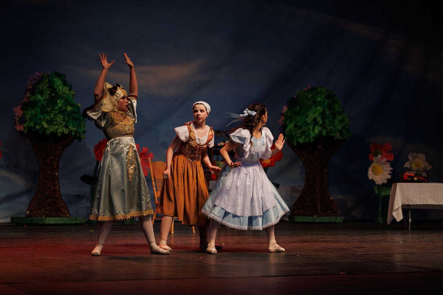 Dancers in rustic costumes perform a folk dance sequence on a dimly lit theatrical stage with scenic backdrop.