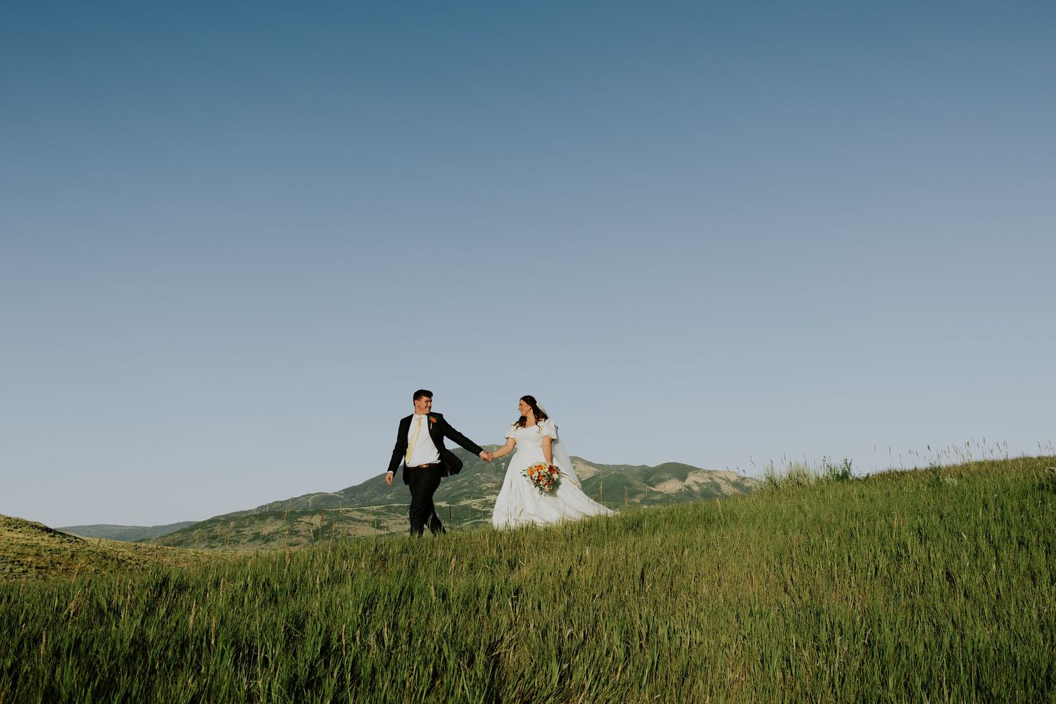 Two figures walk along a scenic green hilltop during a sunny afternoon with blue skies overhead.
