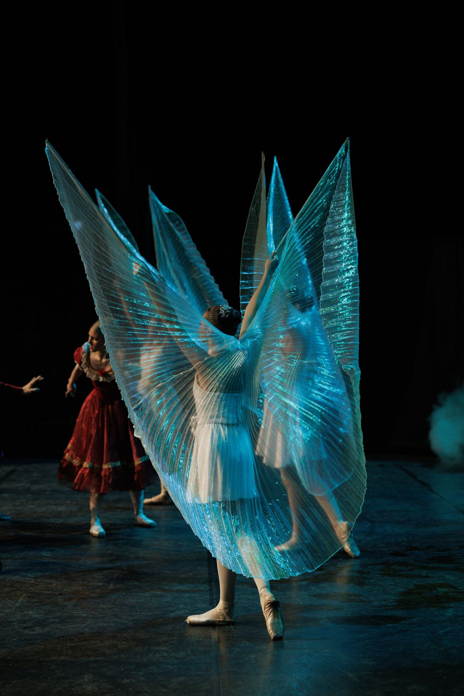 Dancers perform in flowing blue wing-like costumes against a dark stage background.