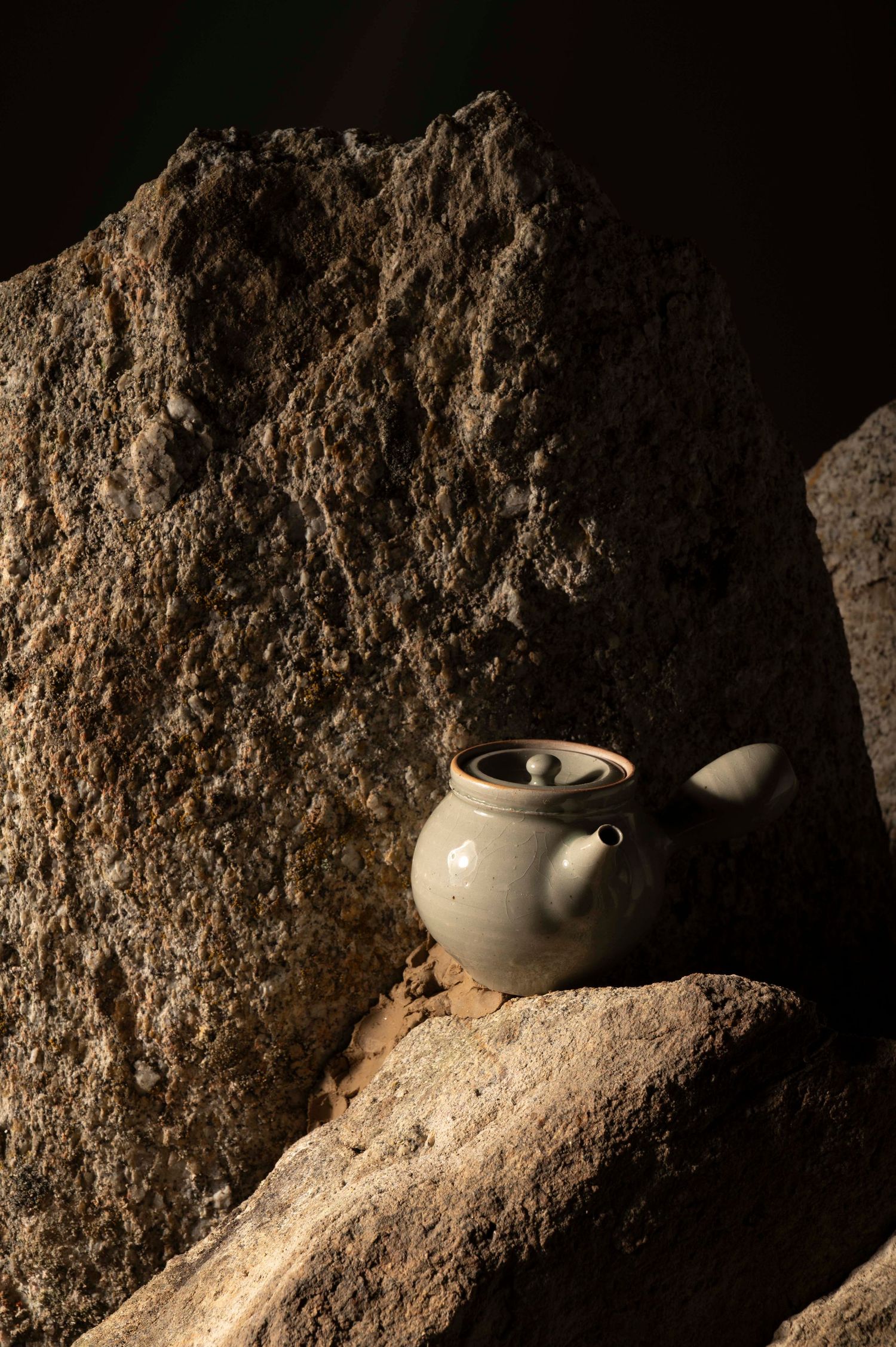 A ceramic teapot sits on a rugged stone surface illuminated by dramatic lighting.