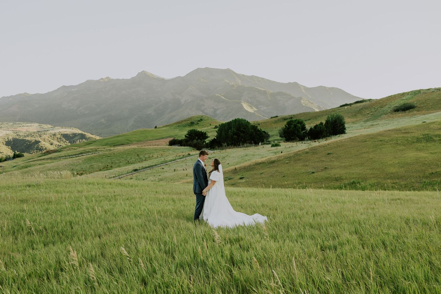 A couple embraces in a flowing white wedding dress against rolling green hills and misty mountains in the background.