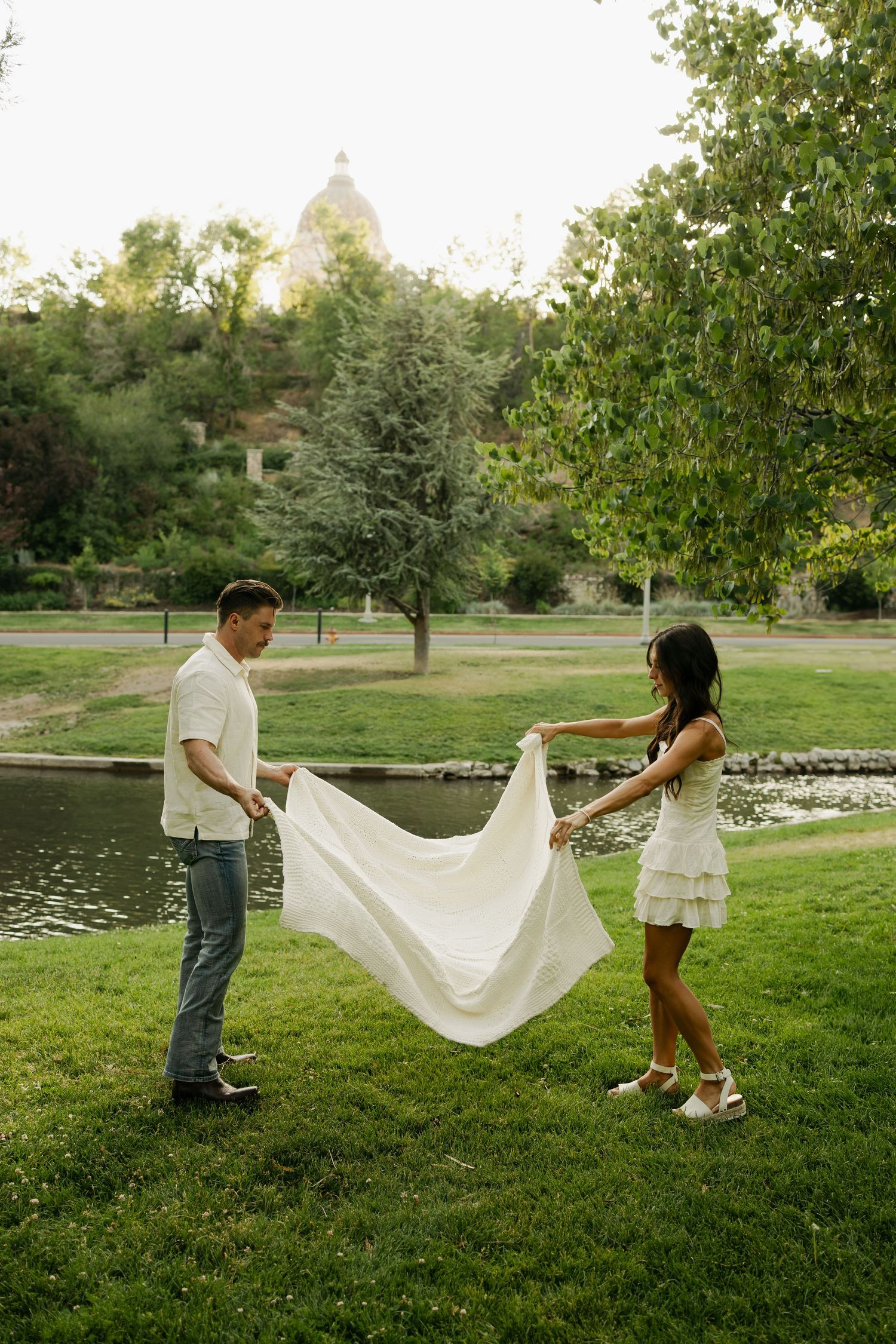 Two people walking through a park holding opposite ends of a flowing white fabric in a romantic outdoor setting.