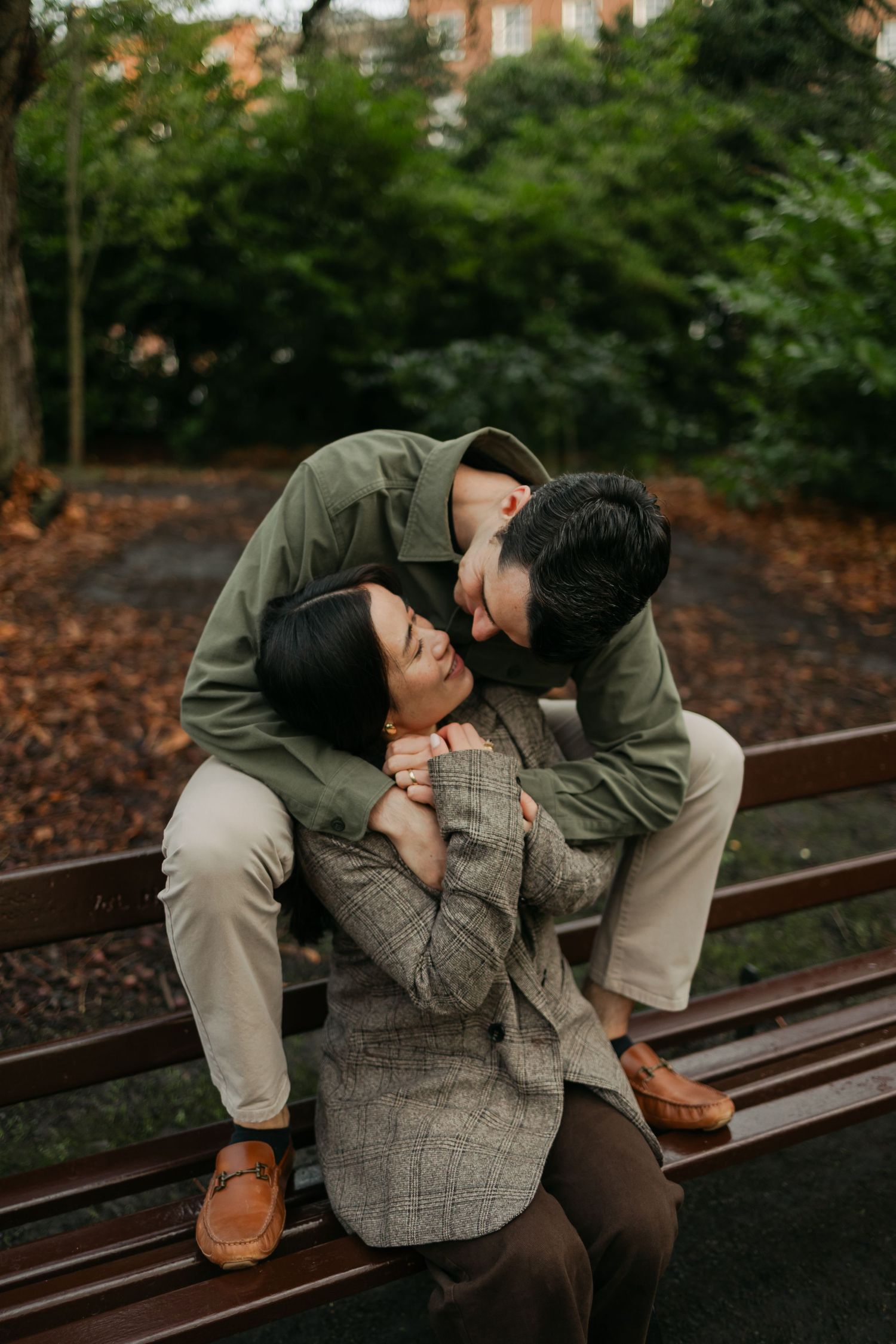 Romantic couple sharing a tender moment on a park bench, man leaning down to kiss woman in plaid blazer.