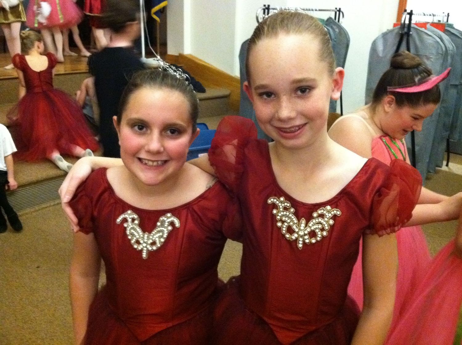 Two young girls in matching red ballet costumes with jeweled bodices smile together backstage at a dance recital.
