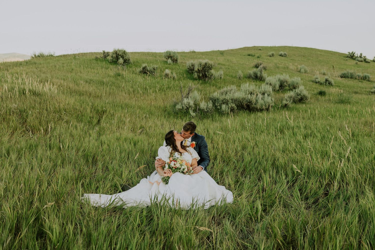 A couple in wedding attire sitting together in a scenic green meadow against rolling hills.