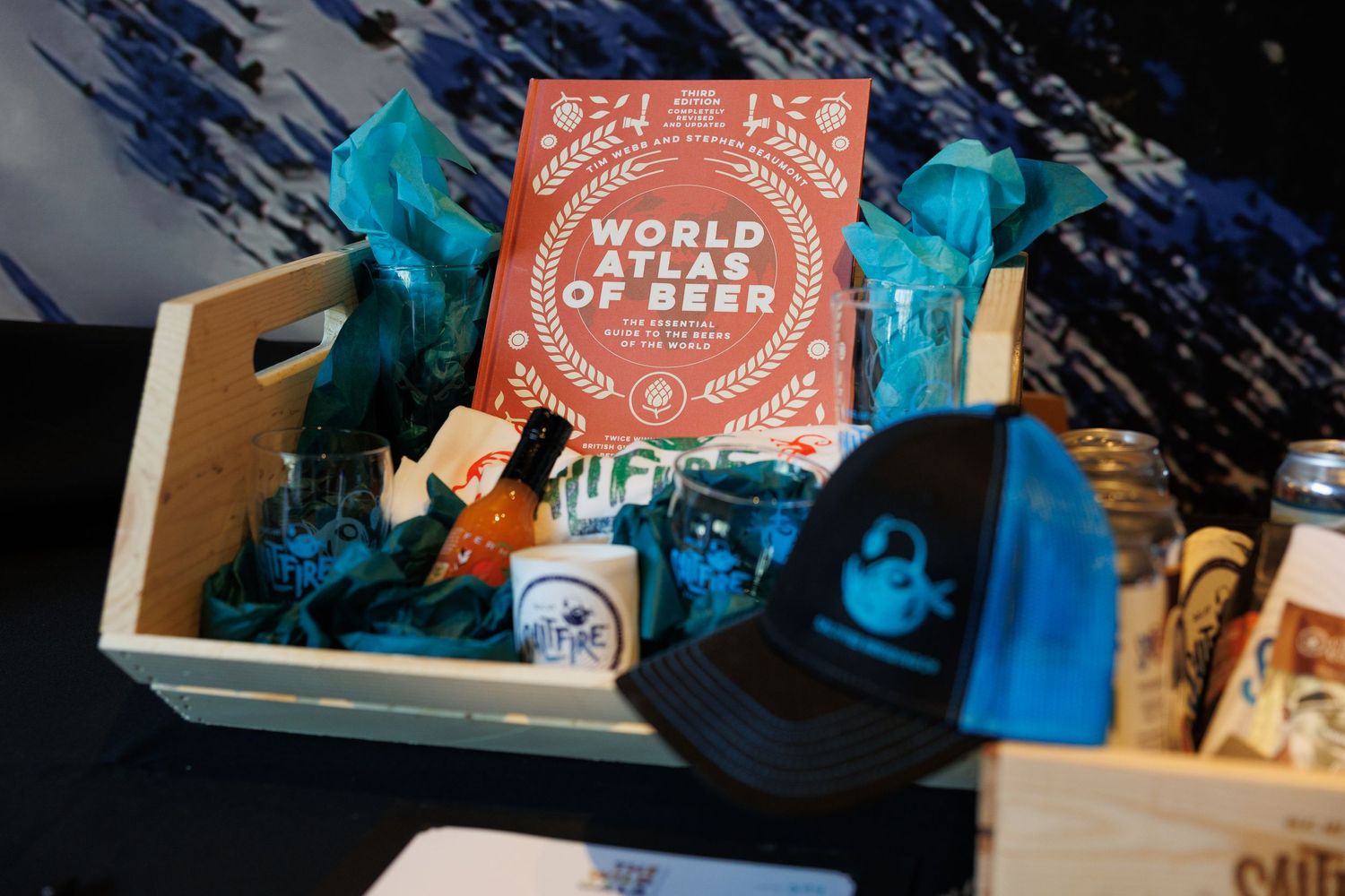 A gift basket featuring World Atlas of Beer book, blue tissue paper, hat, and assorted beer accessories in a wooden crate.