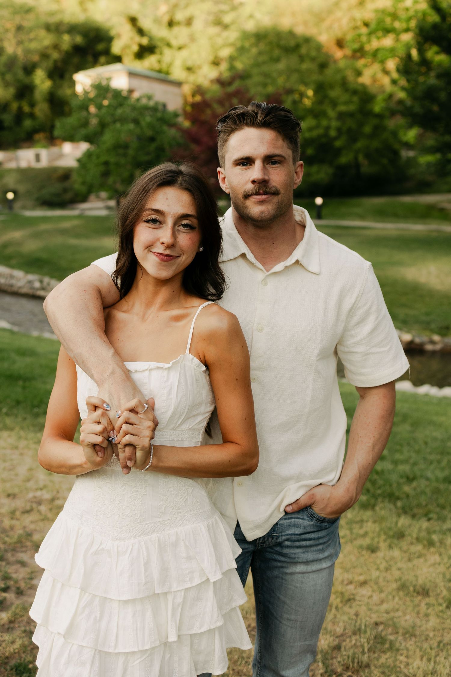 A couple in white summer outfits poses together on a grassy lawn during golden hour with trees in the background.