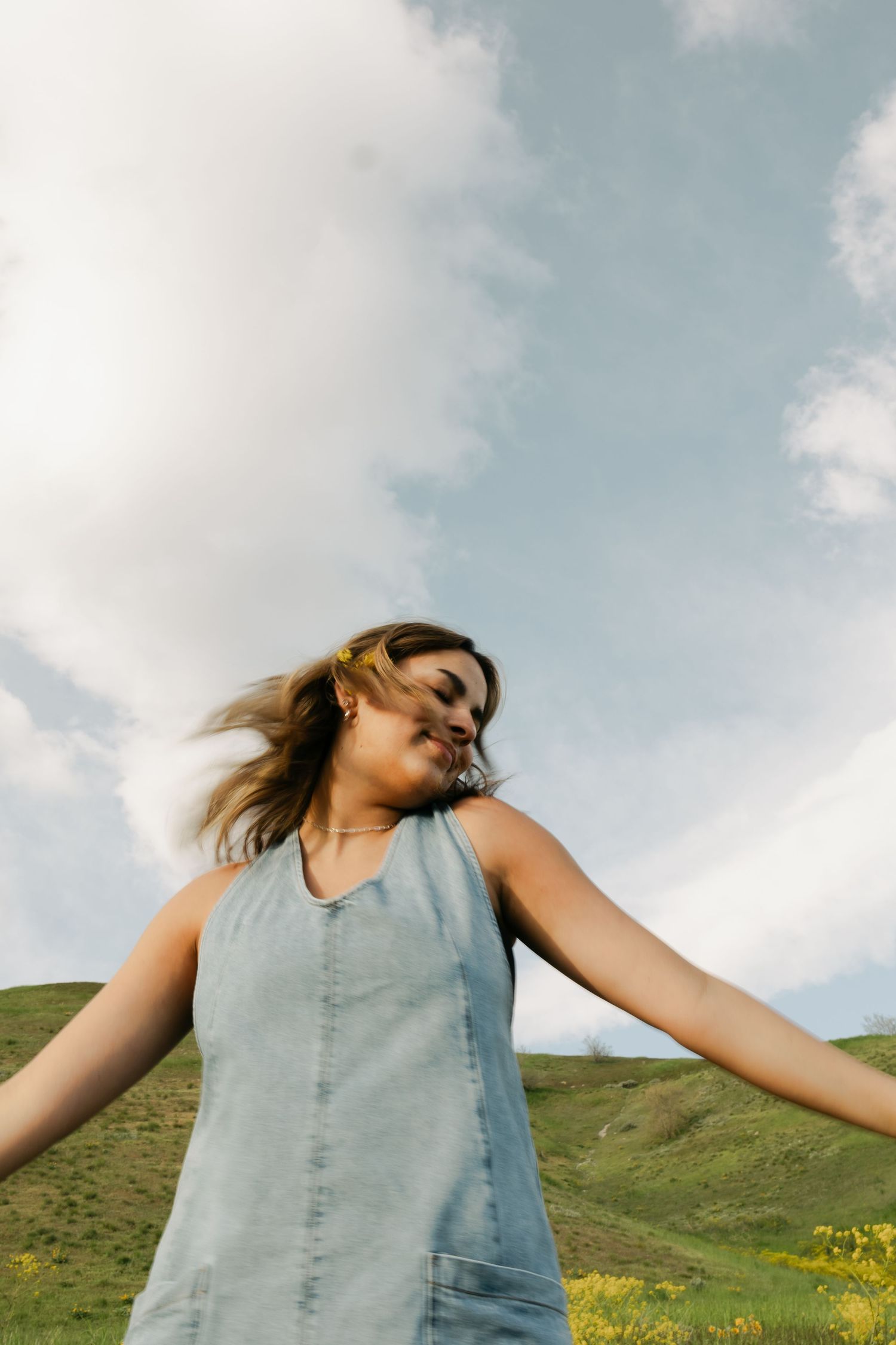 Person in denim dress twirling against blue sky and green hillside.