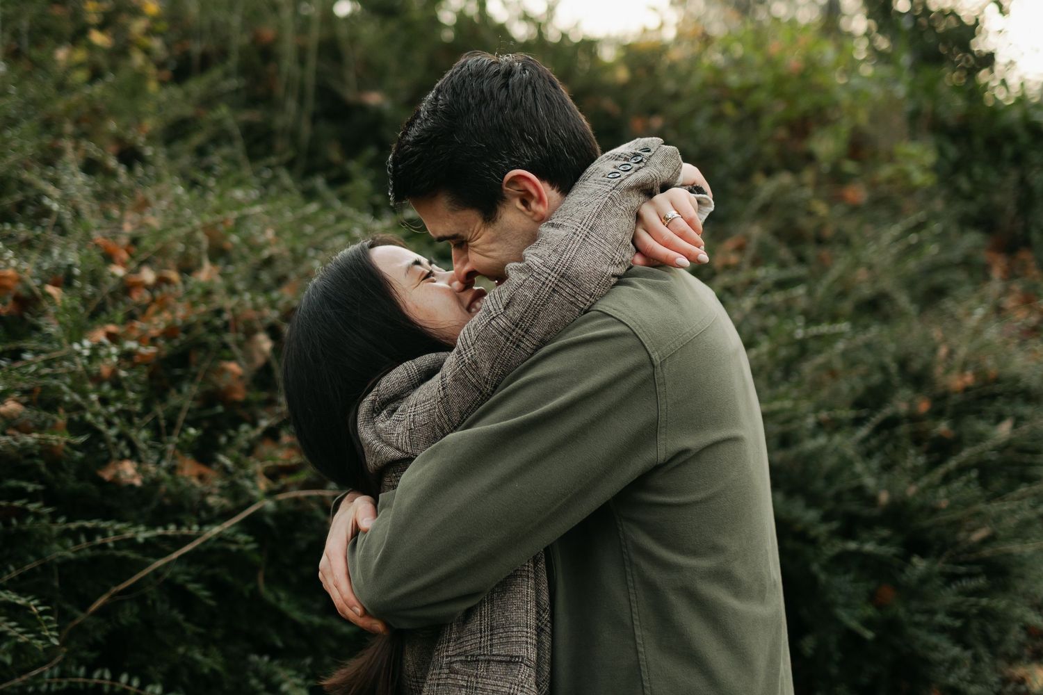 A couple embraces warmly outdoors, surrounded by lush green foliage in an autumn setting.