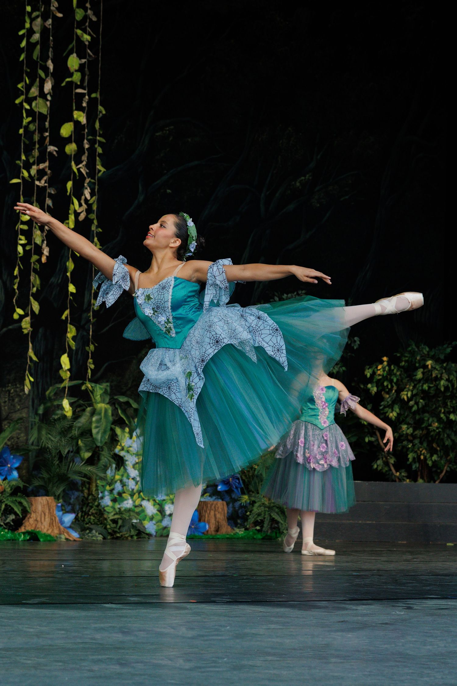 Ballet dancers in turquoise tutus perform graceful poses on stage with vine decorations in the background.