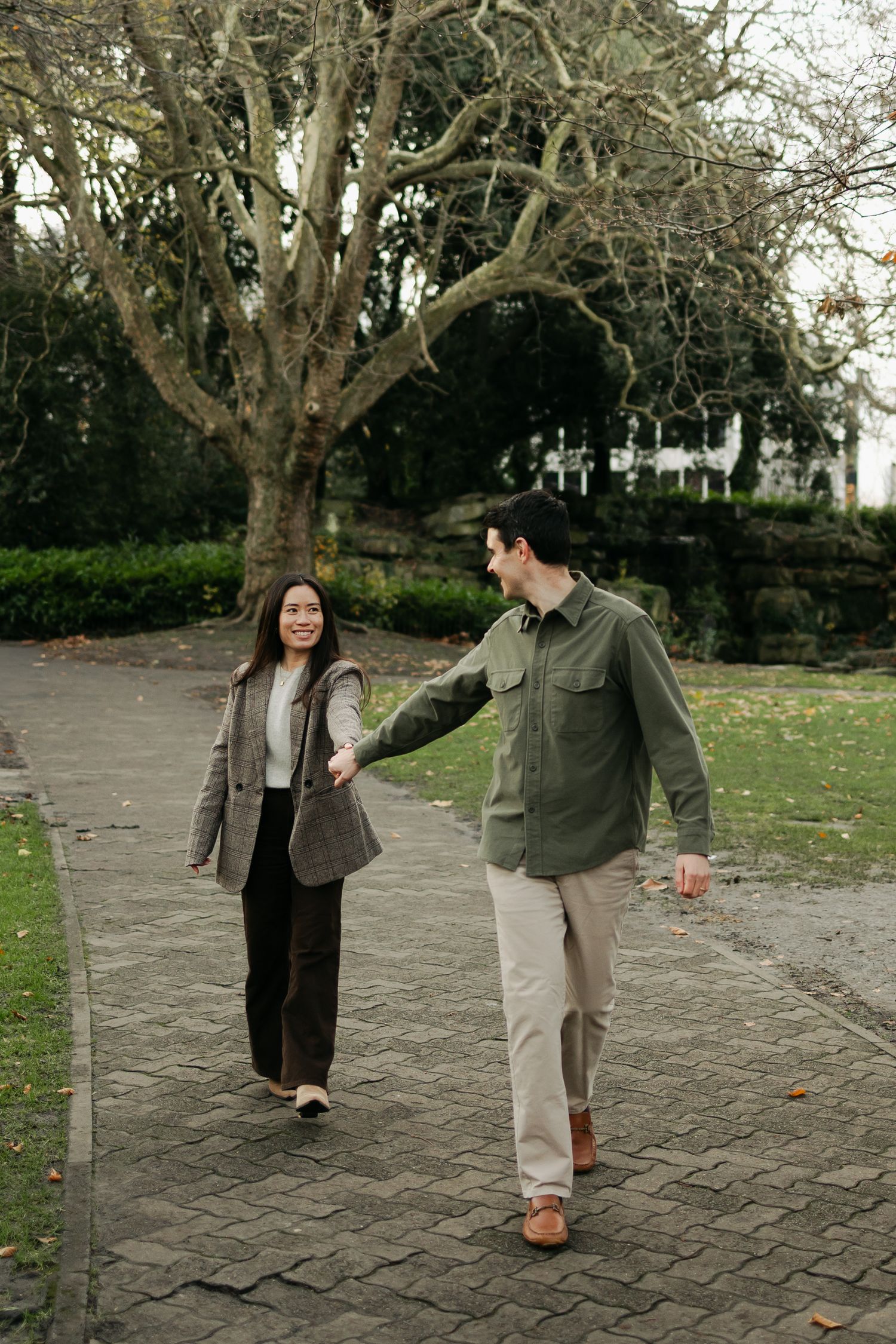 Happy couple holding hands and smiling while strolling along a paved park path with bare winter trees.
