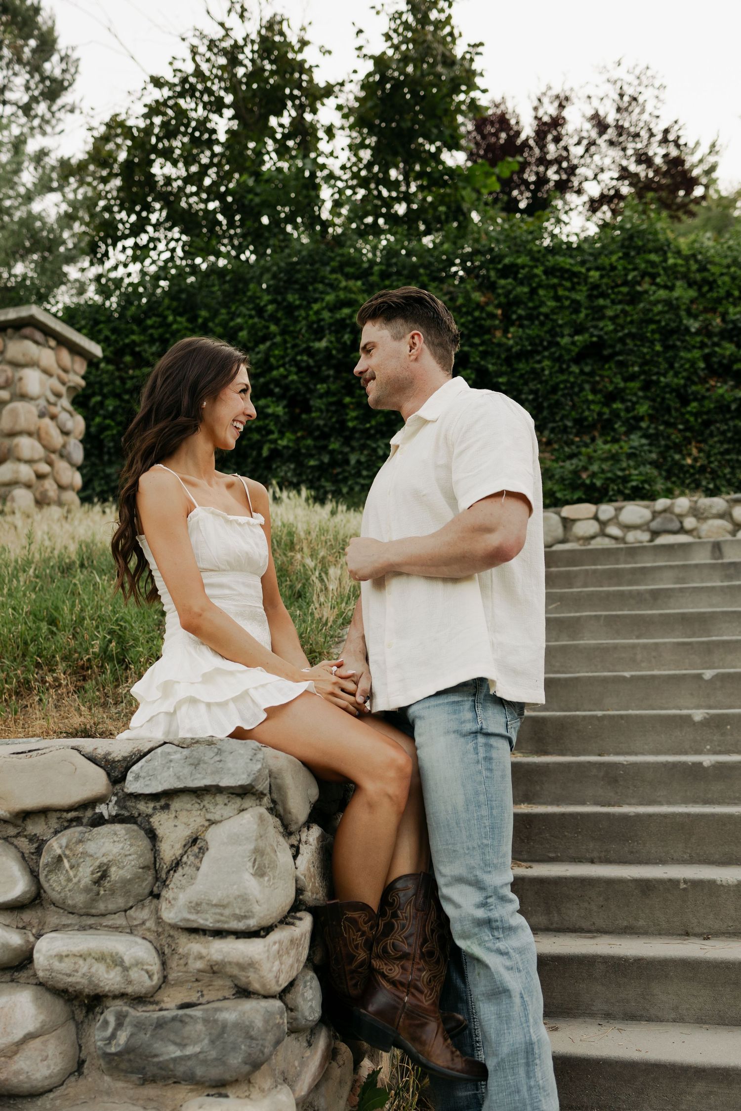 A romantic couple share a joyful moment together on stone steps surrounded by greenery on a summer evening.