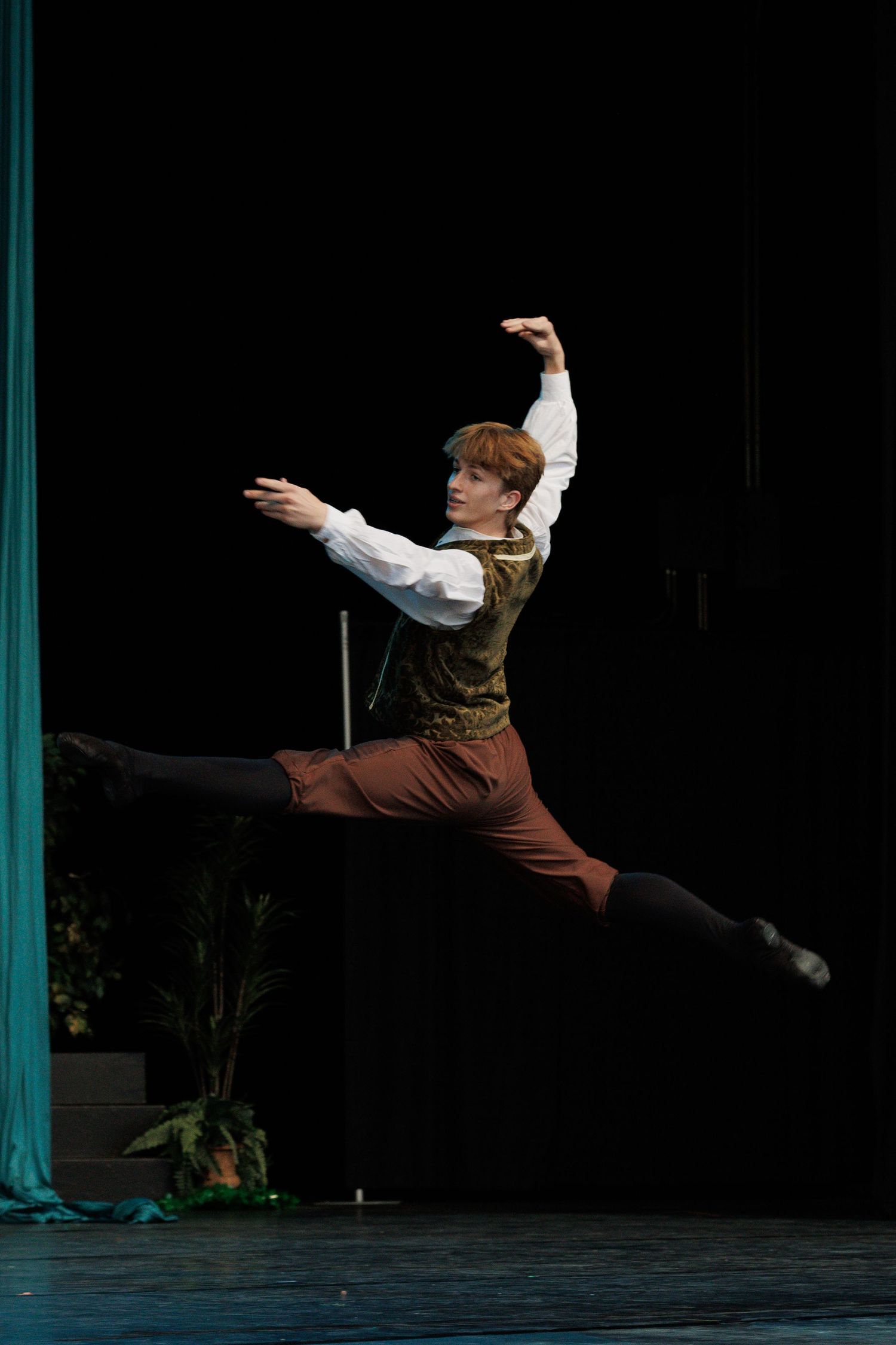 Male ballet dancer performs dramatic leap sequence in brown costume against dark stage with turquoise accents.