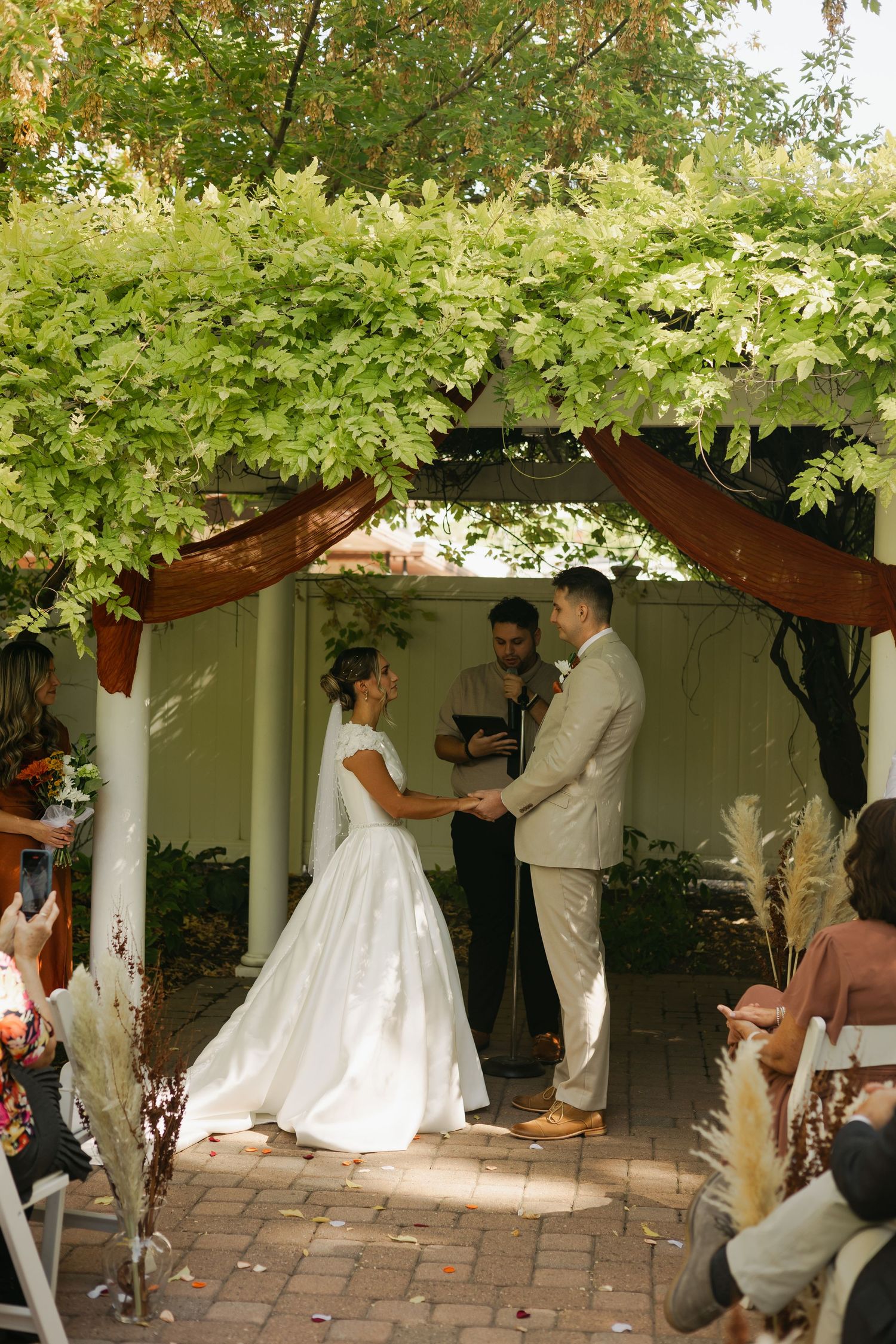 Wedding ceremony under a vine-covered arch with guests seated on either side.